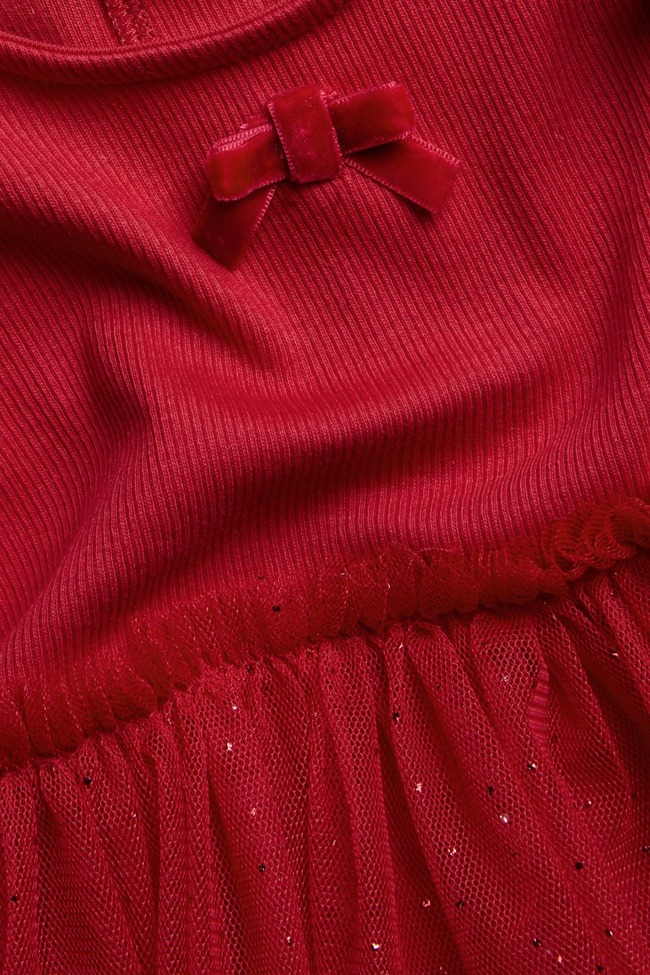 Zbliżenie na czerwoną sukienkę dla niemowląt z prążkowaną górą, aksamitną kokardą i brokatową tiulową spódnicą.