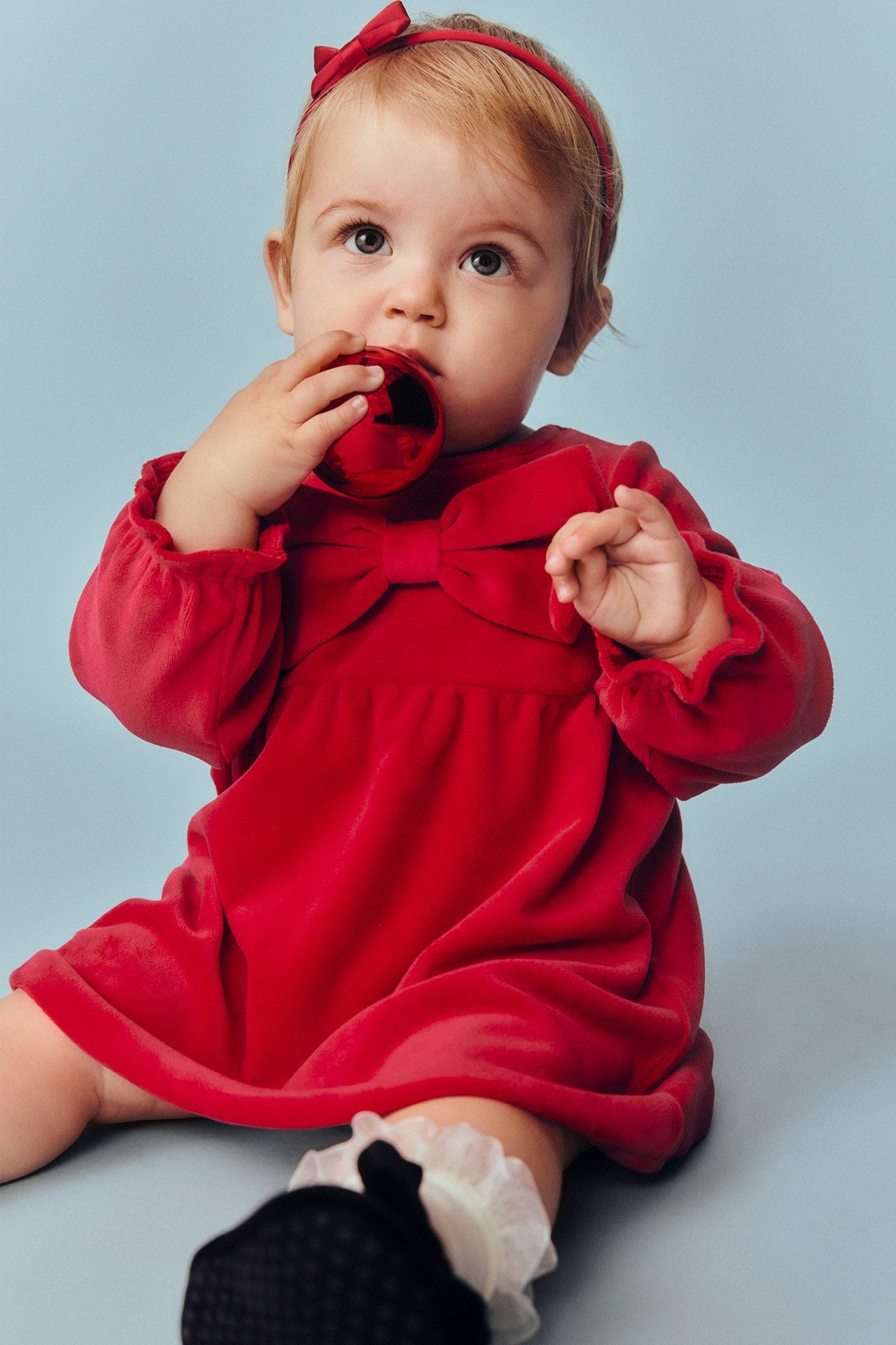 Edestäpäin vauva punaisessa samettimekossa rusetilla, punainen hiuspanta, pitää punaista palloa.