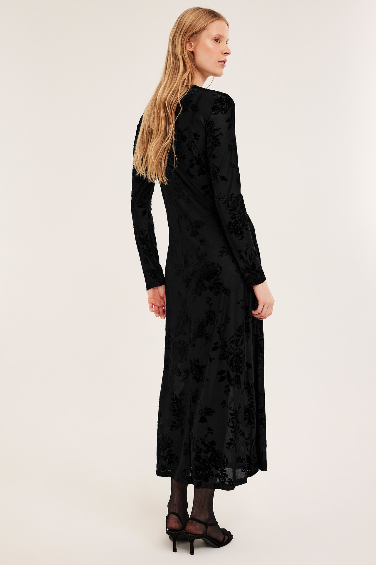 Trekvart ryggutsikt av kvinne i svart langermet blomstret midikjole med svarte strømpebukser og sandaler.