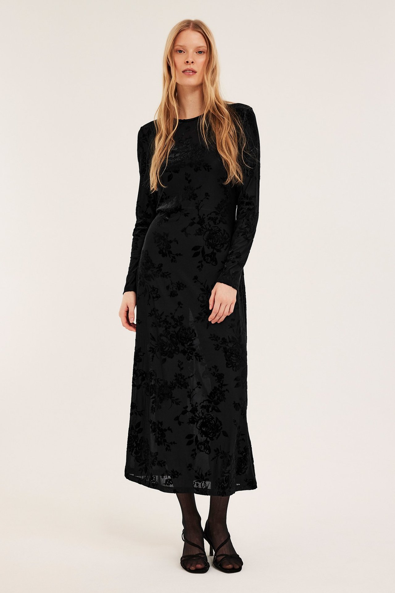 Forfra, kvinne i sort blomstret flokktrykt kjole, sorte strømpebukser og sorte sandaler med høye hæler.