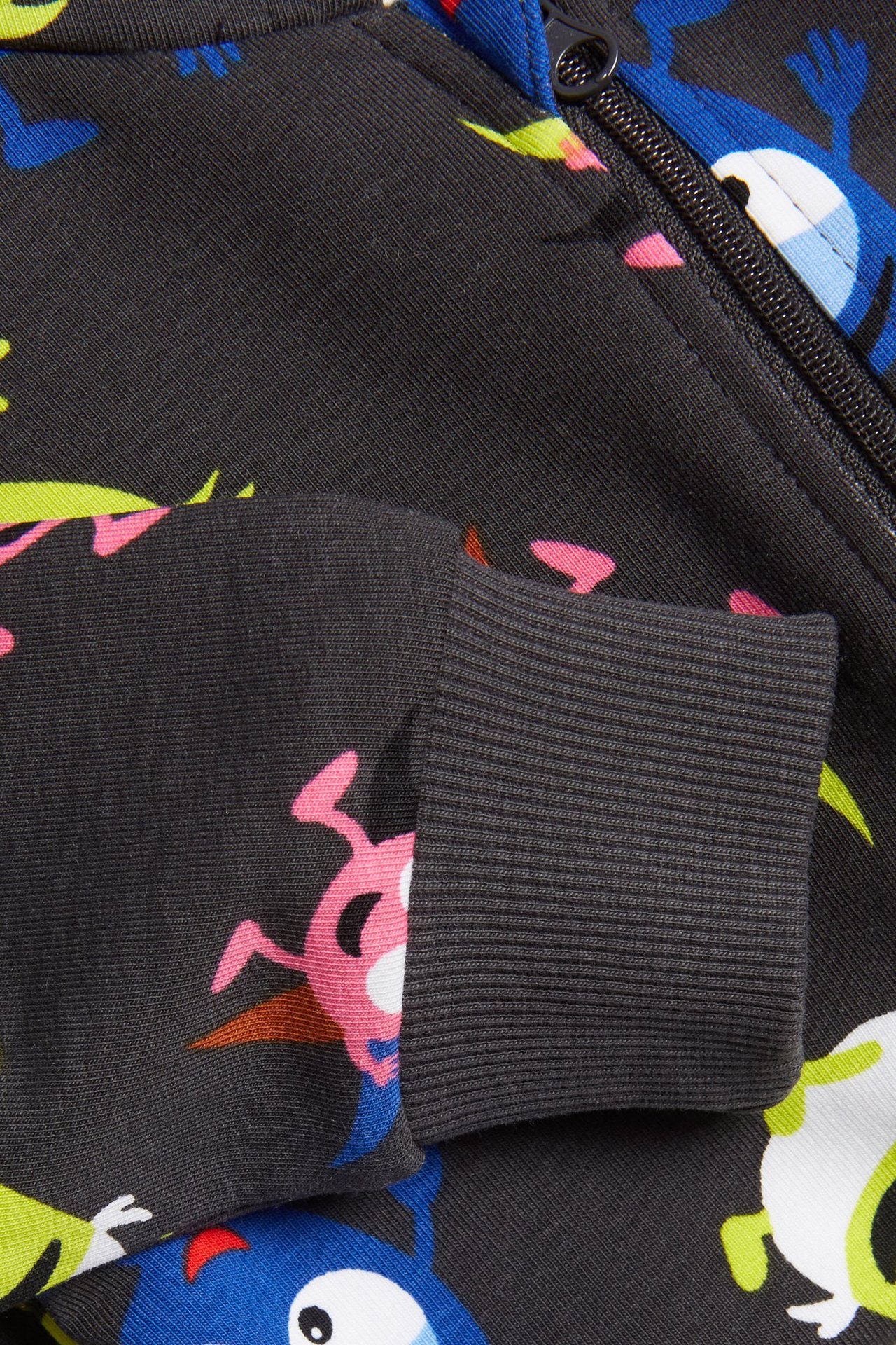 Zbliżenie na ciemnoszary wzorzysty kombinezon niemowlęcy z kolorowym nadrukiem Babblarna, ściągaczem i czarnym zamkiem.