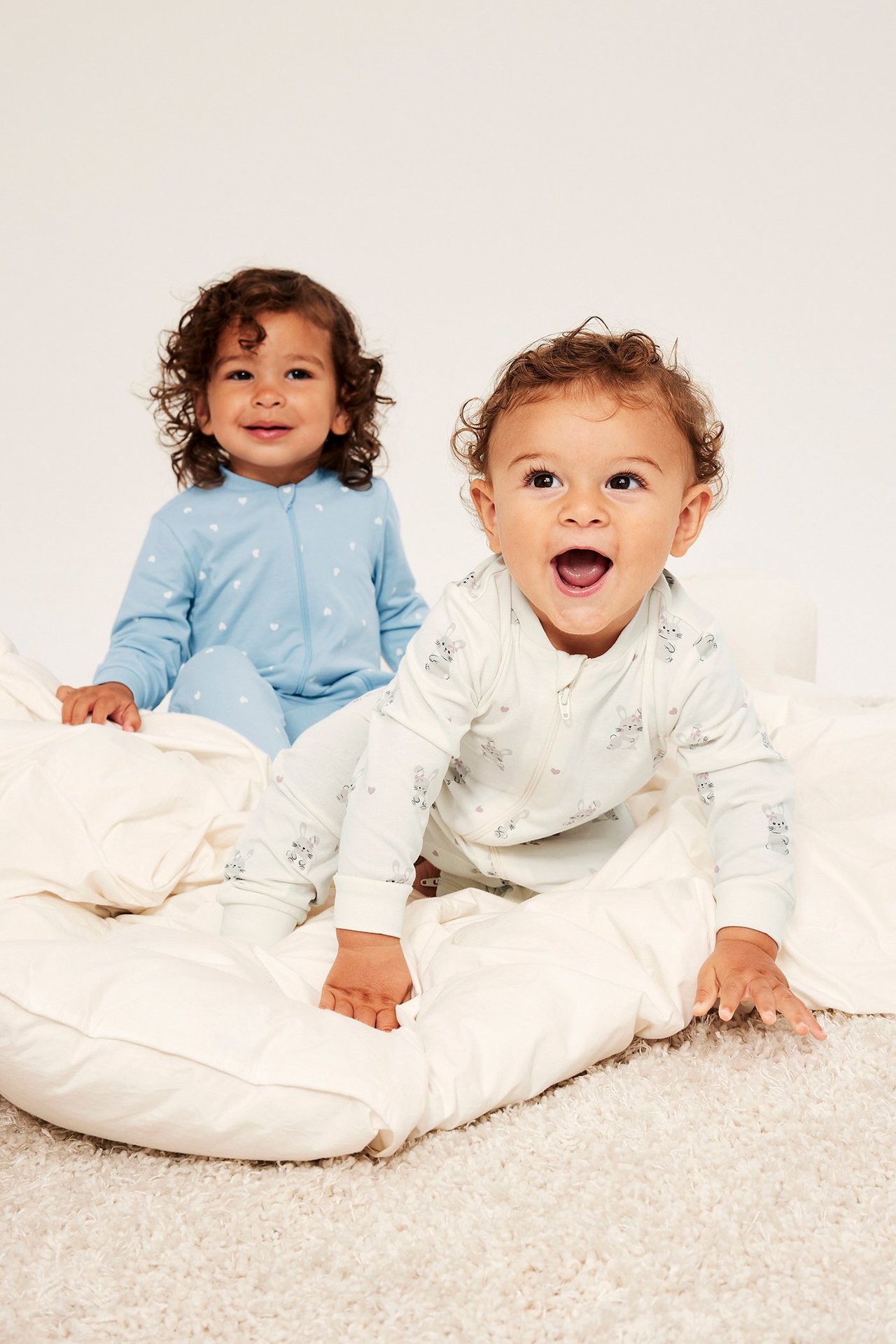 Forfra. Baby i off-white kaninpysjamas med glidelås; annen baby i lys blå hjertepysjamas.