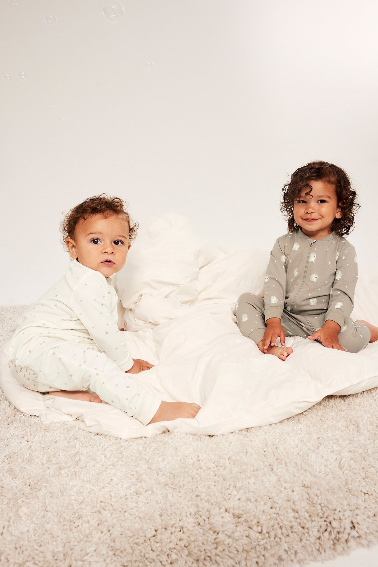 Kaksi vauvaa etupuolelta kuvattuna. Toisella harmaanbeige siiliröykkiöpuku vetoketjulla, toisella vaaleanbeige pilkullinen röykkiöpuku.