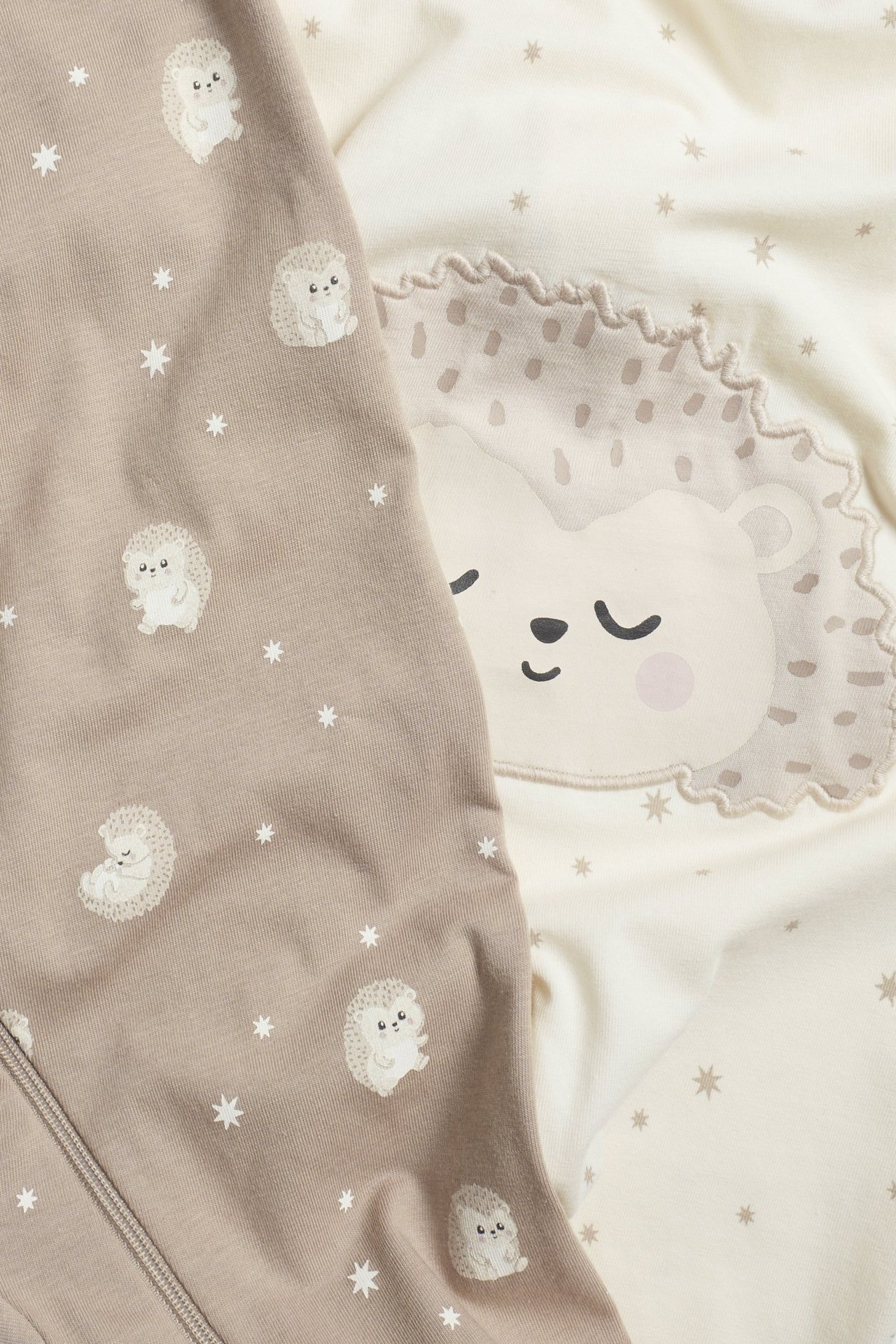 Zbliżenie dwóch piżam dla niemowląt: beżowa w jeże i ecru z dużą twarzą jeża.