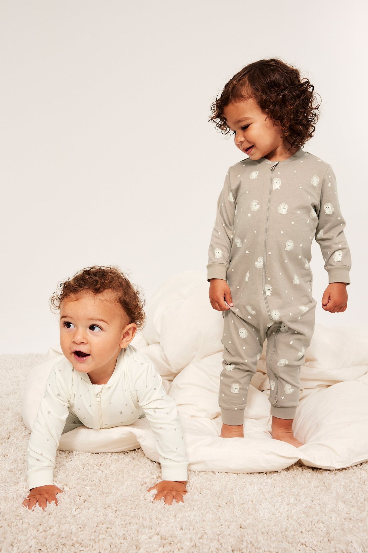Widok z przodu: Dziecko w beżowym pajacyku z jeżami i zamkiem. Drugie dziecko w jasnobeżowym w gwiazdki.