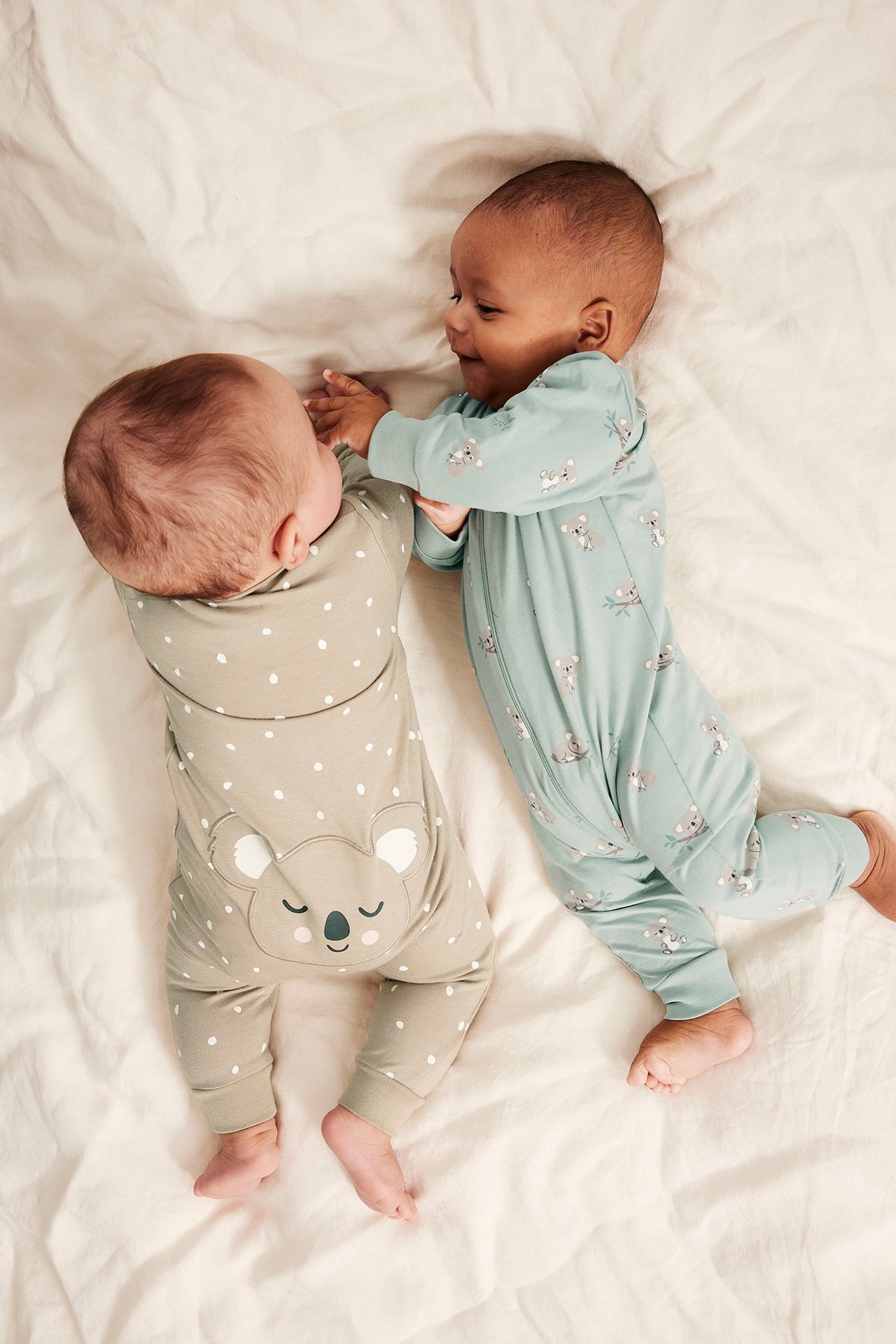 Två bebisar i en ljusgrön pyjamas med koalatryck och en ljusbeige prickig pyjamas.