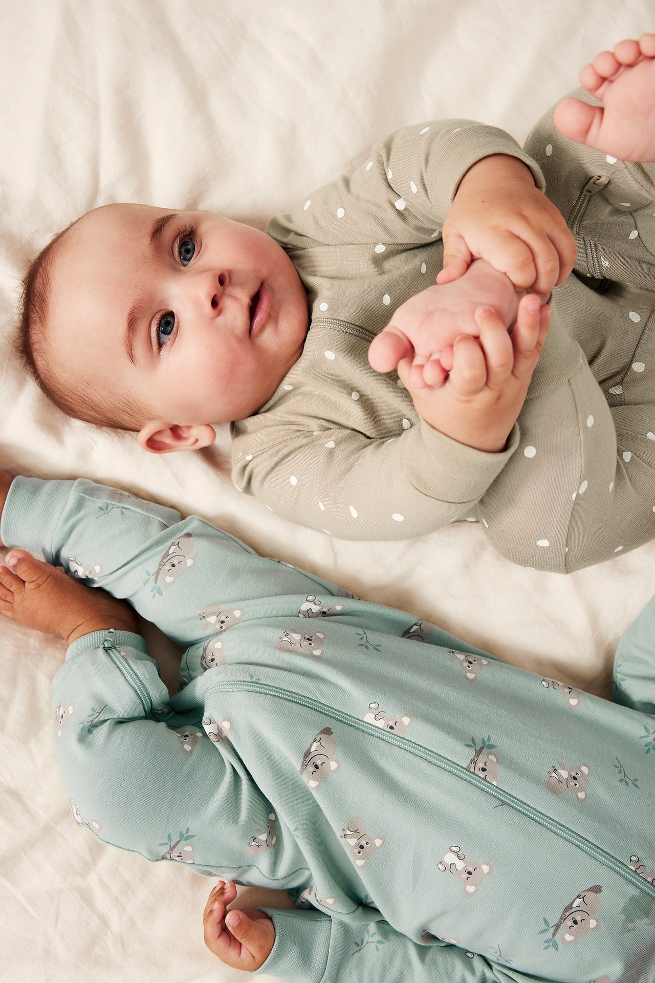 Dwoje niemowląt w pajacykach: jasnozielony w kropki i jasnoniebieski z nadrukiem koali, oba z zamkami.