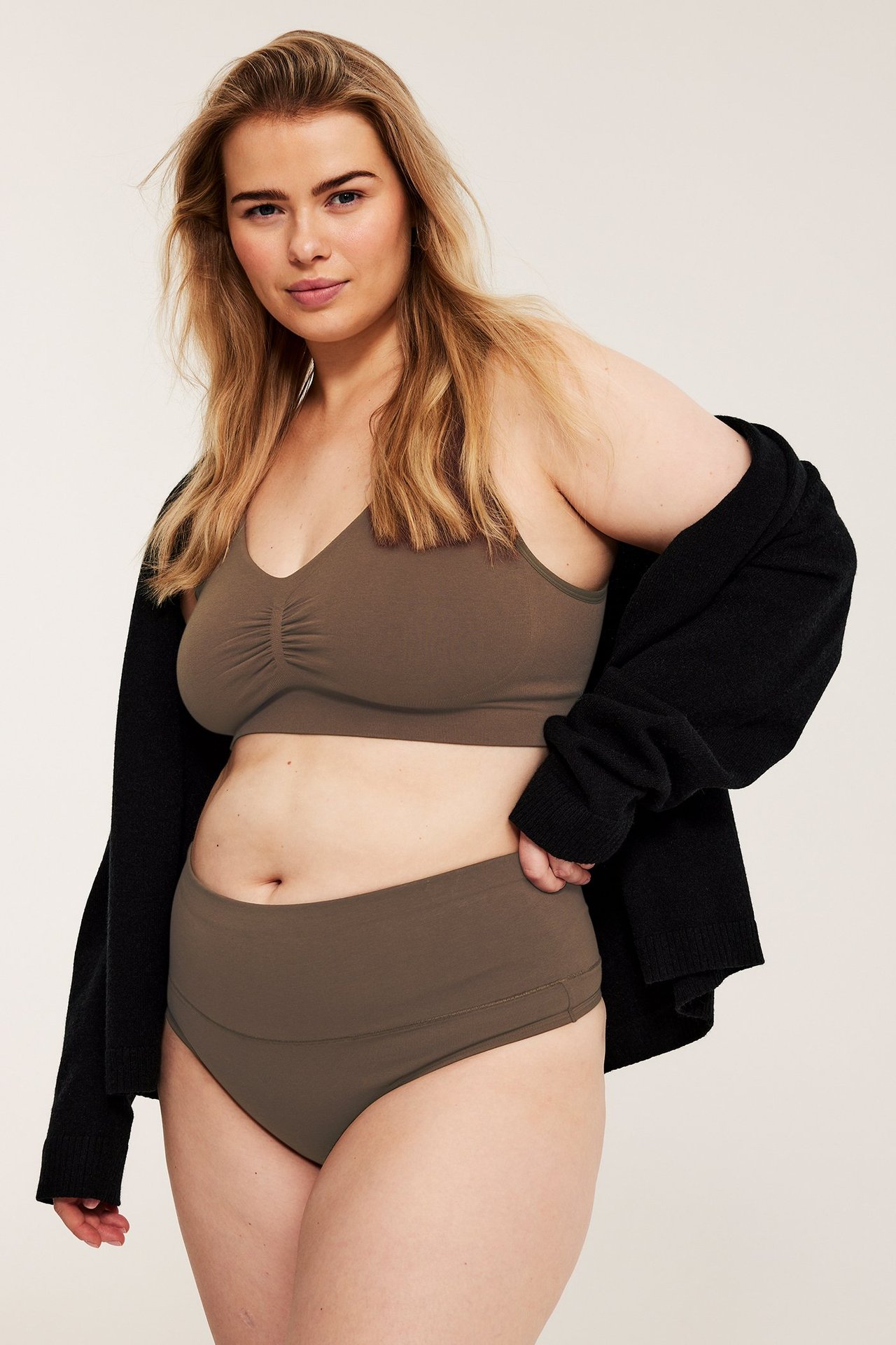 Frontvy av kvinna i mörkbrun bomullstrosa och matchande bralette, med svart kofta draperad över axlarna.