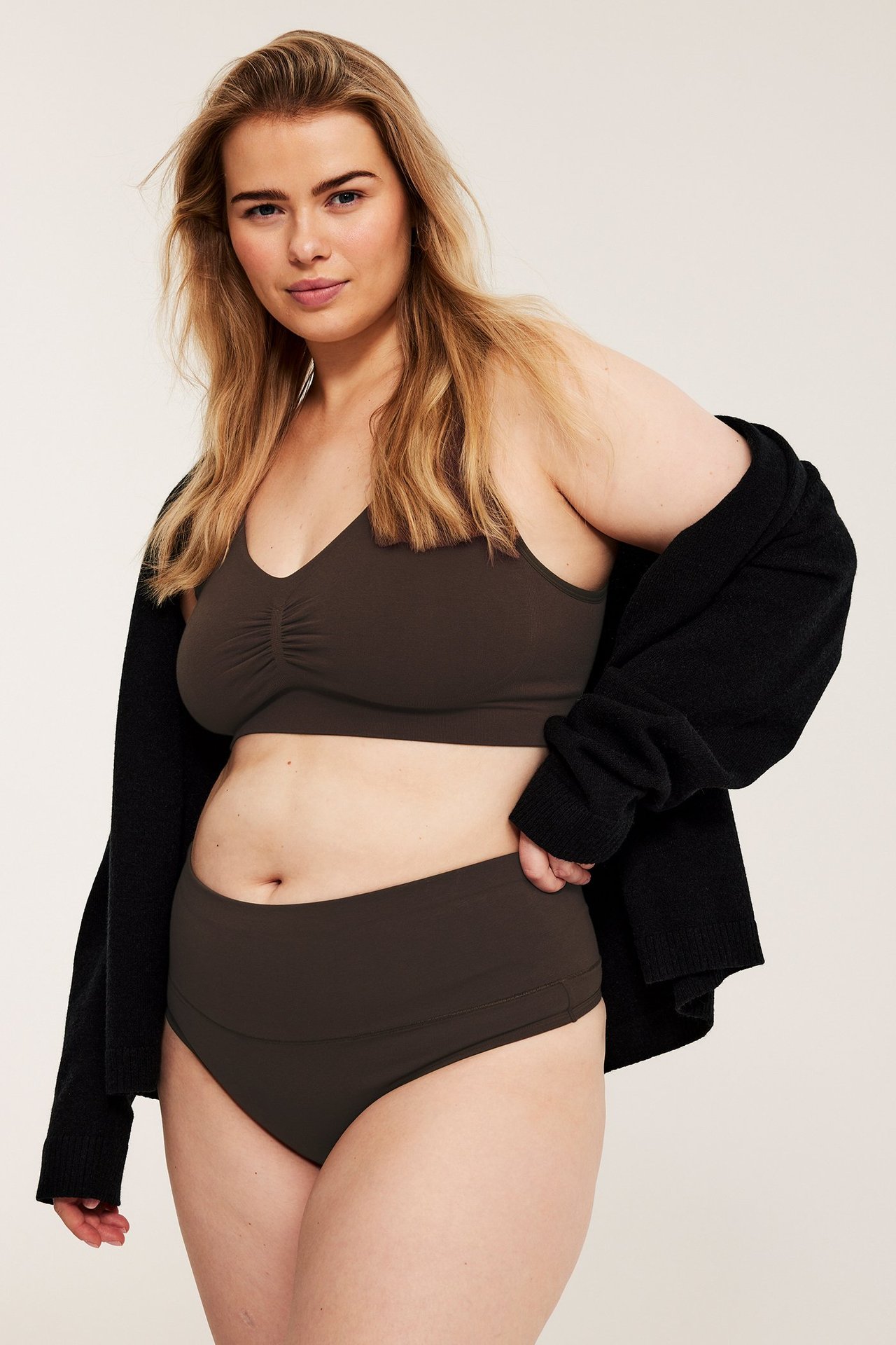 Frontvy av en kvinna i mörkbrun hög bomullsstringtrosa, matchande bralette och svart kofta.