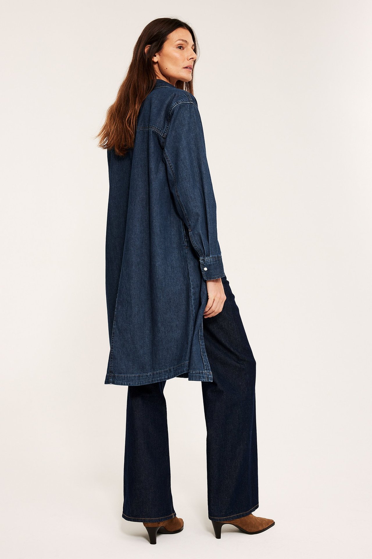 Bokiem kobieta w długiej, ciemnoniebieskiej koszulowej sukience jeansowej, ciemnych jeansach i brązowych botkach.