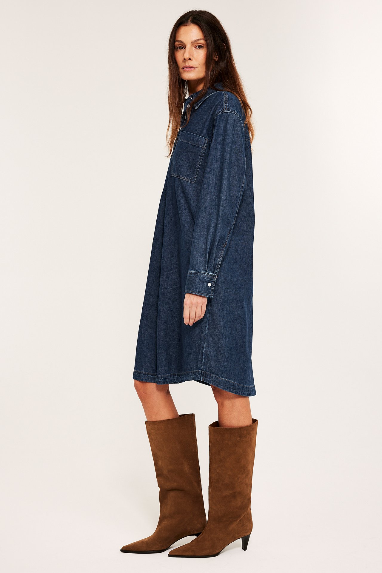Sidevisning av kvinne i lang mørk denimskjortekjole og brune semskede knestøvler.