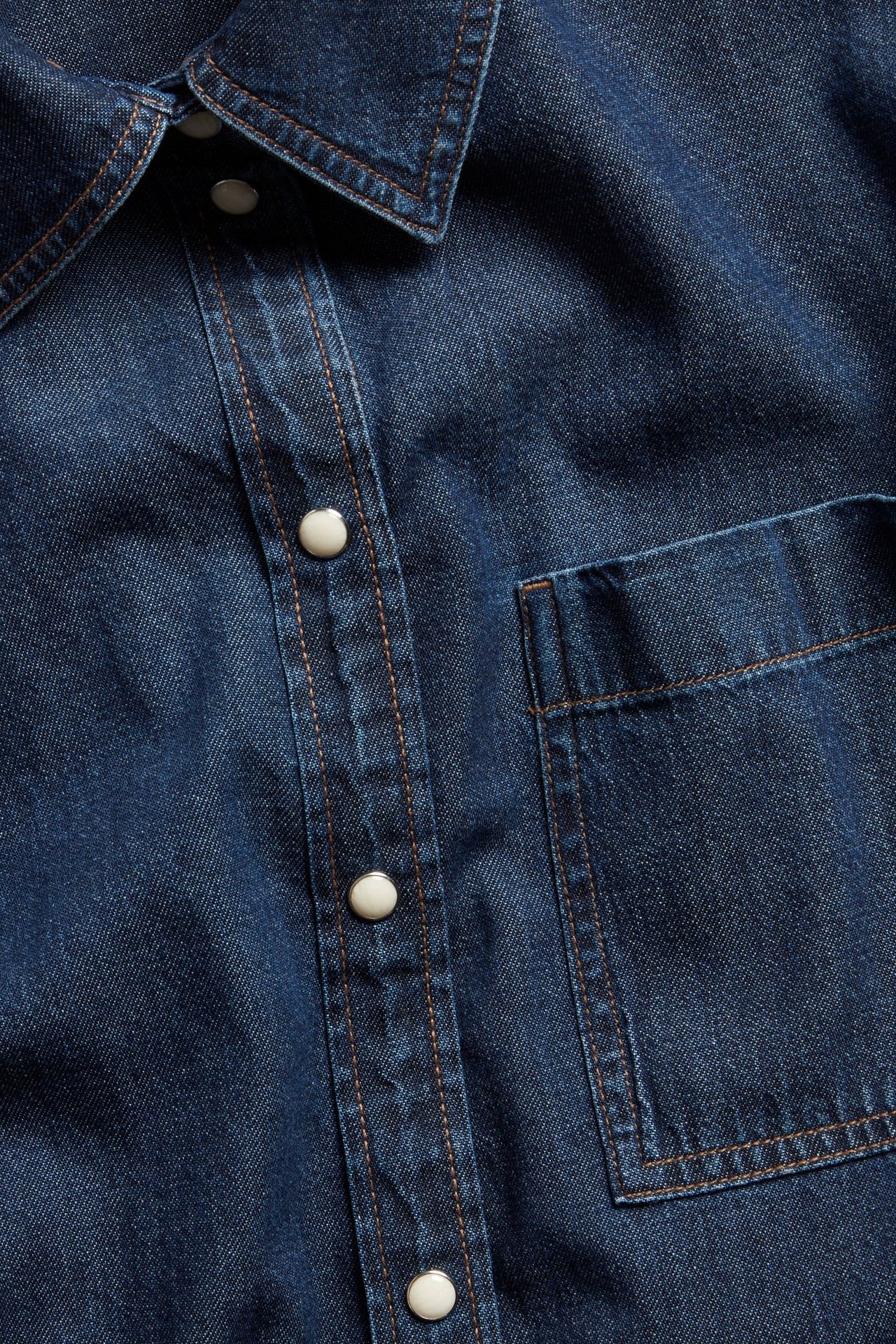Zbliżenie ciemnoniebieskiej sukienki jeansowej z białymi zatrzaskami, kołnierzem i kontrastowymi przeszyciami.