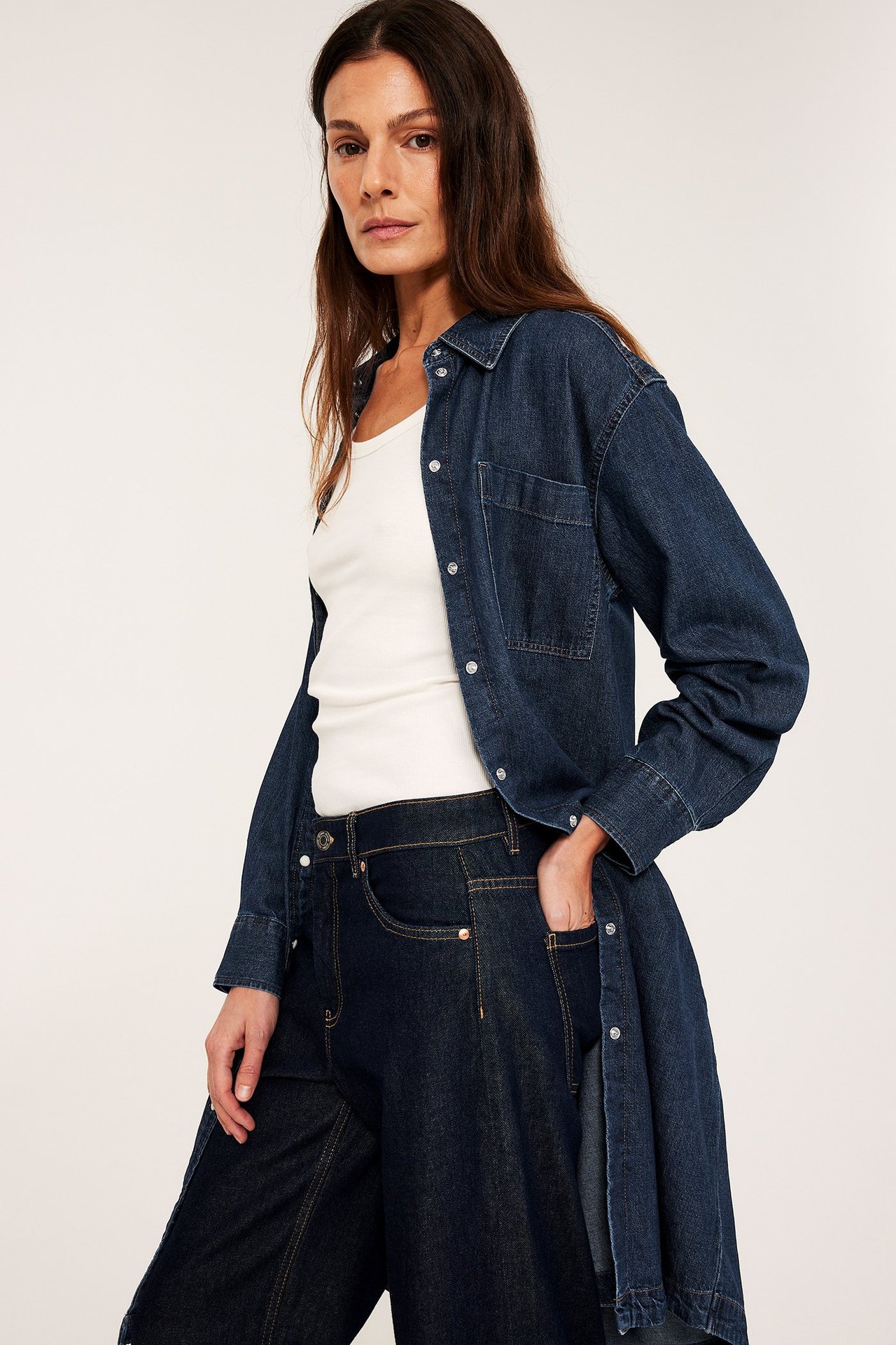 Forfra: Kvinne i lang mørk denim skjortekjole, åpen over hvit ribbet tanktopp og mørke jeans.