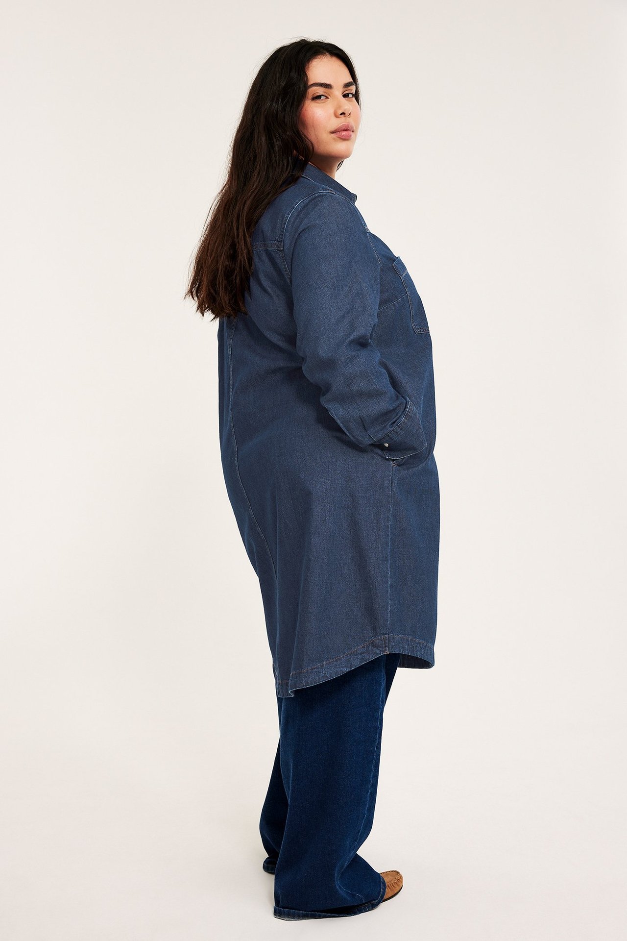 Sivukuva naisesta tummassa farkkupaitamekossa sivutaskuilla, yllään siniset farkut ja ruskeat kengät.