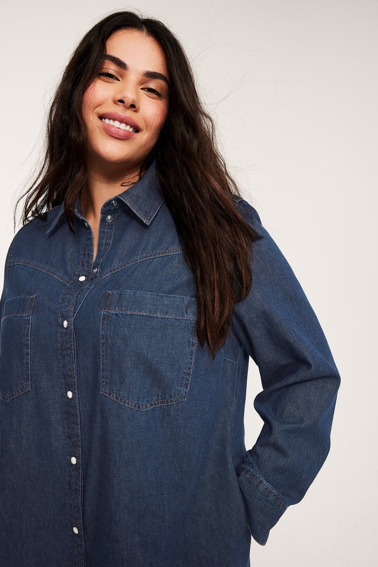 Forfra: kvinne i en mørkeblå dongerikjole med hvite trykknapper og brystlommer.