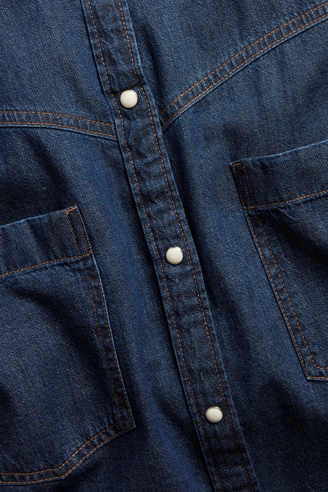 Zbliżenie na ciemnoniebieską sukienkę dżinsową z białymi napami, kontrastowymi szwami i kieszeniami na piersi.