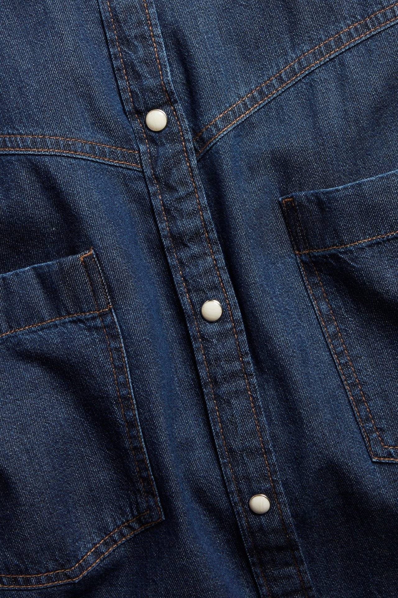 Nærbilde av en mørk denimskjortekjole med hvite trykknapper, kontrastsømmer og brystlommer.
