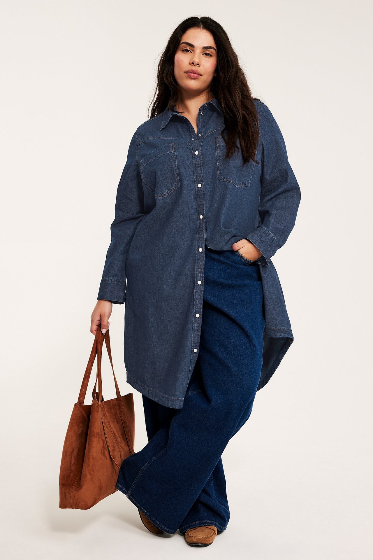 Forfra: Kvinne i mørkeblå bomulls denimskjortekjole, vide mørkeblå jeans, holder en brun semsket veske.