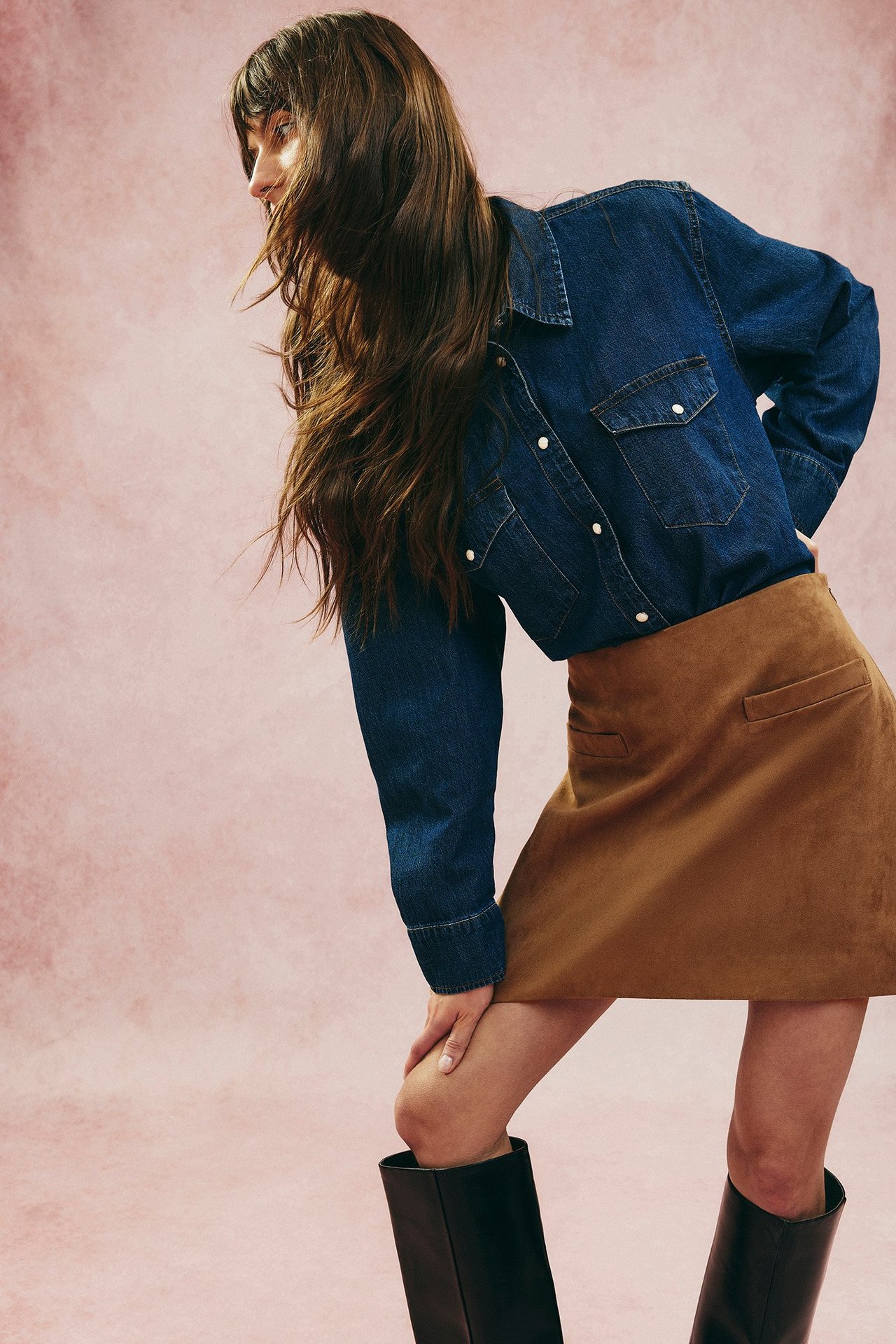 Sidovy av kvinna i mörk denim jeansskjorta med vita knappar, brun minikjol och svarta stövlar.