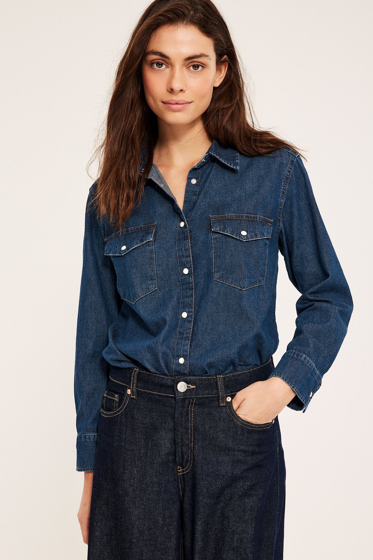 Forfra, kvinne i en mørk jeansskjorte med hvite knapper, brystlommer, gjemt i mørke jeans.