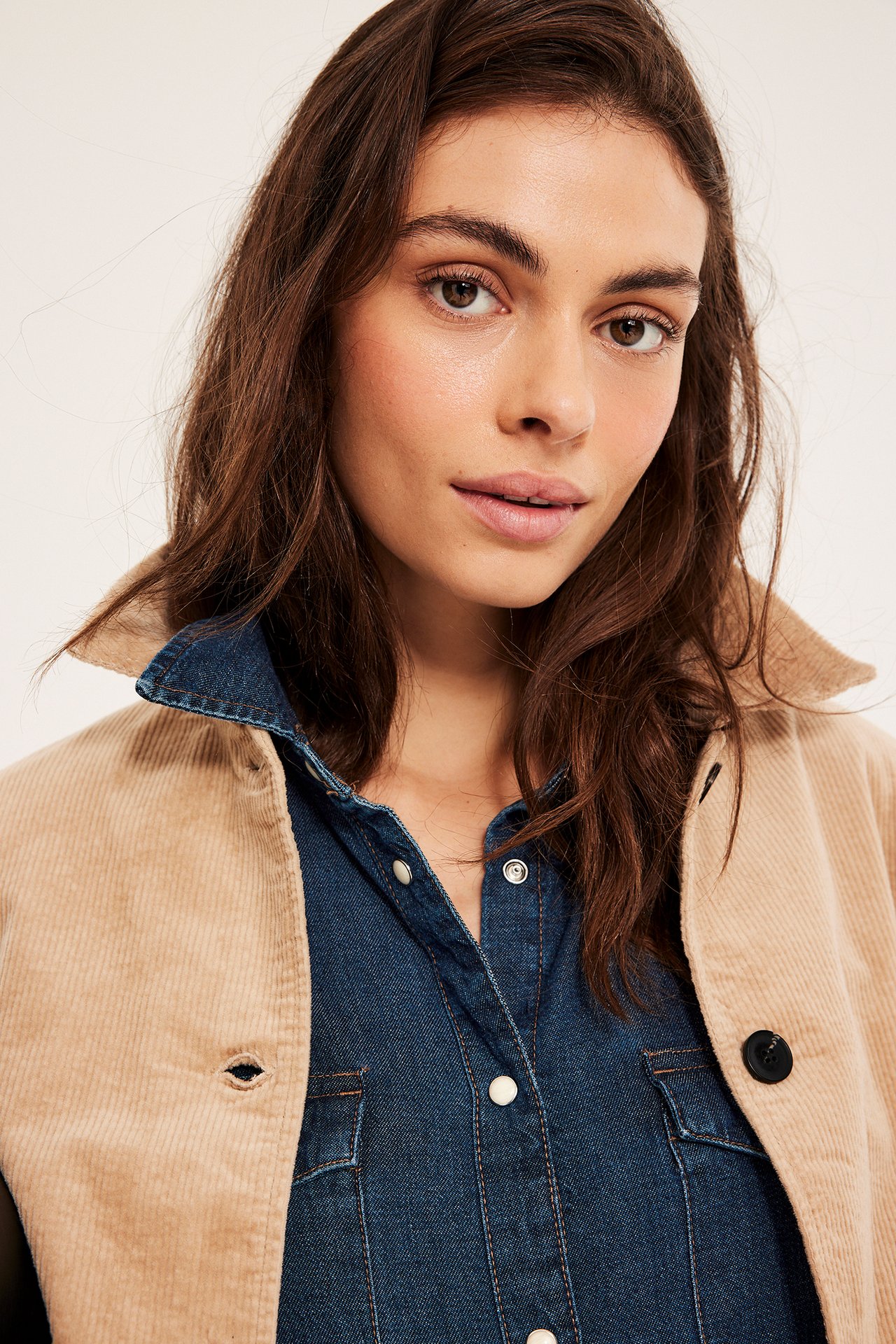 Nærbilde av kvinne i mørkeblå dongeriskjorte med trykknapper, under en beige cordfløyel overskjorte.