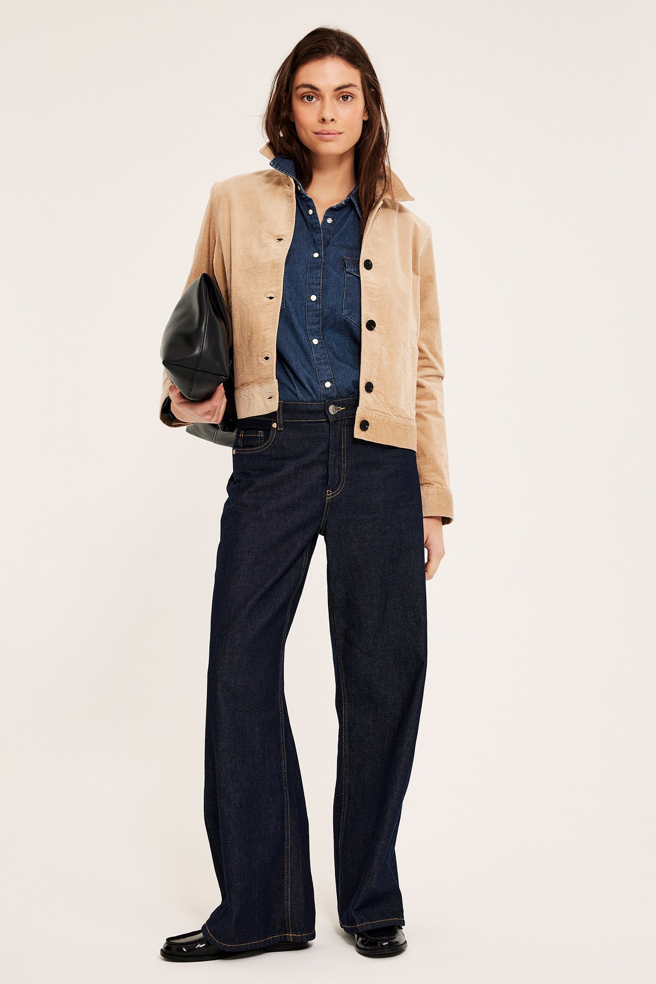 Forfra: Kvinne i mørkblå dongeriskjorte, beige jakke, vide jeans, holder svart håndveske.