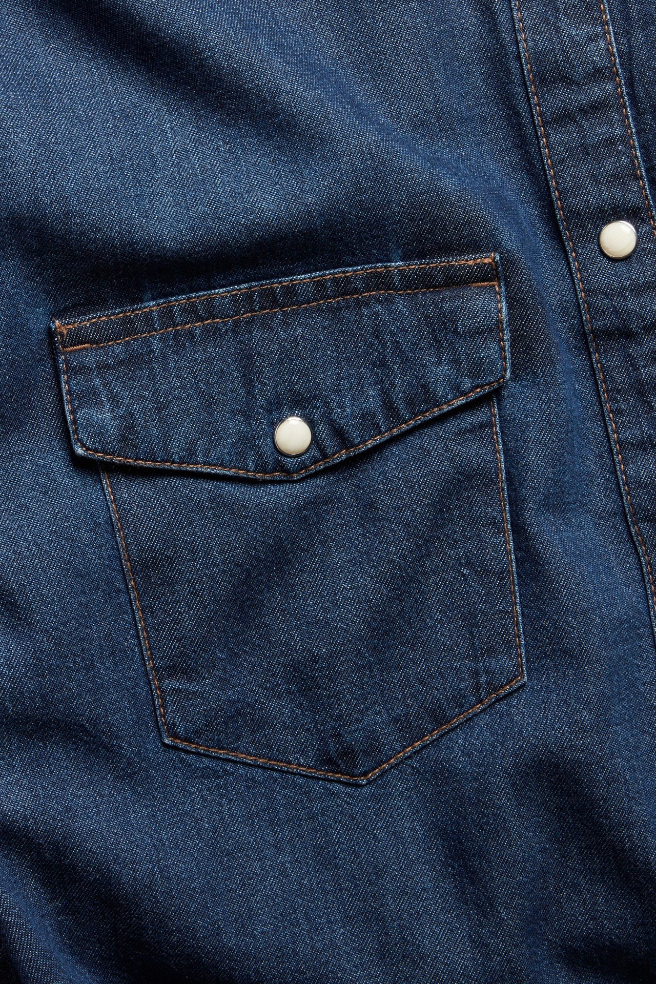 Nærbilde av en mørk denim bomullsskjorte med brystlomme, klaff og hvite trykknapper.