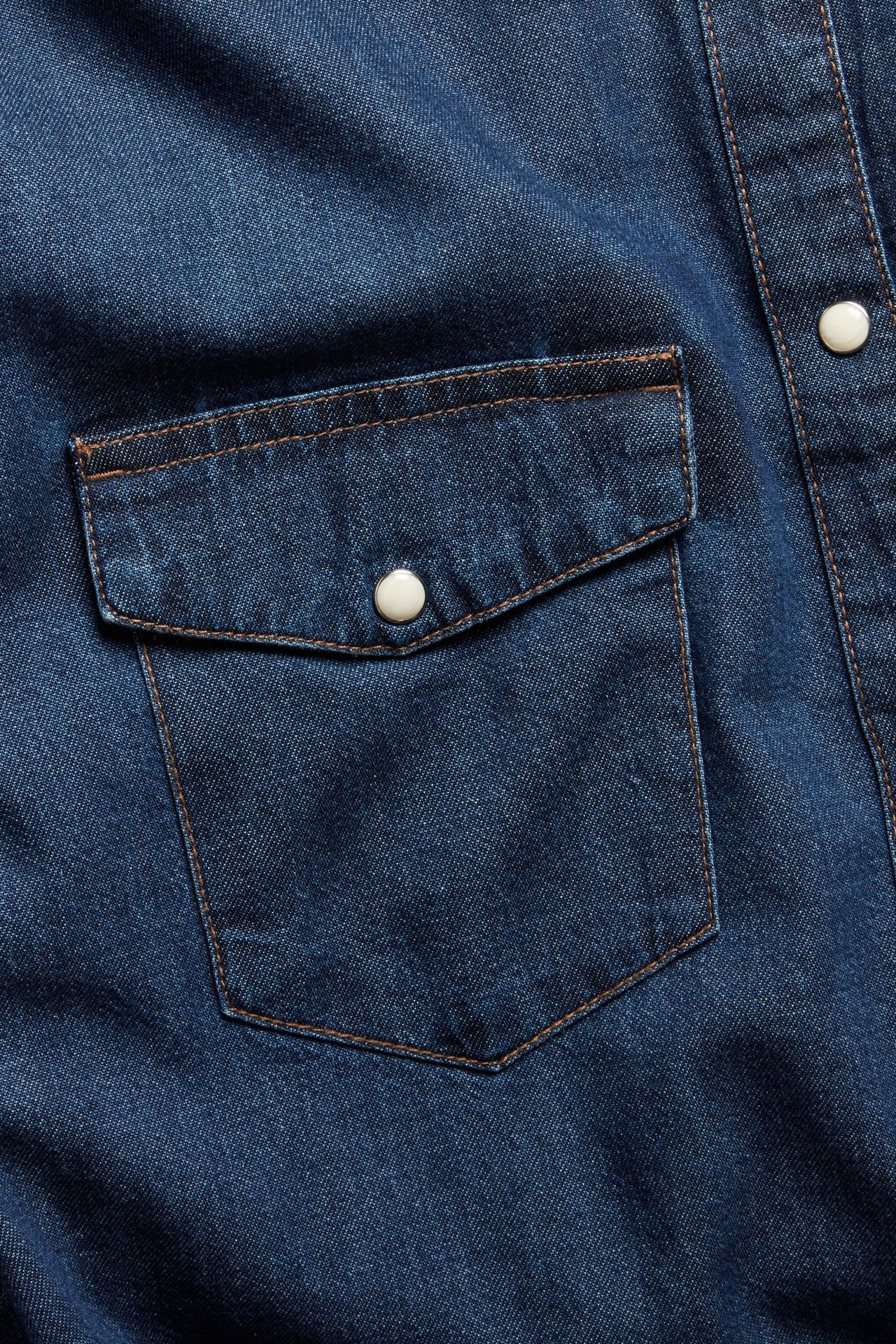 Zbliżenie ciemnogranatowej koszuli jeansowej z bawełny z kieszenią, klapą i białymi napami.