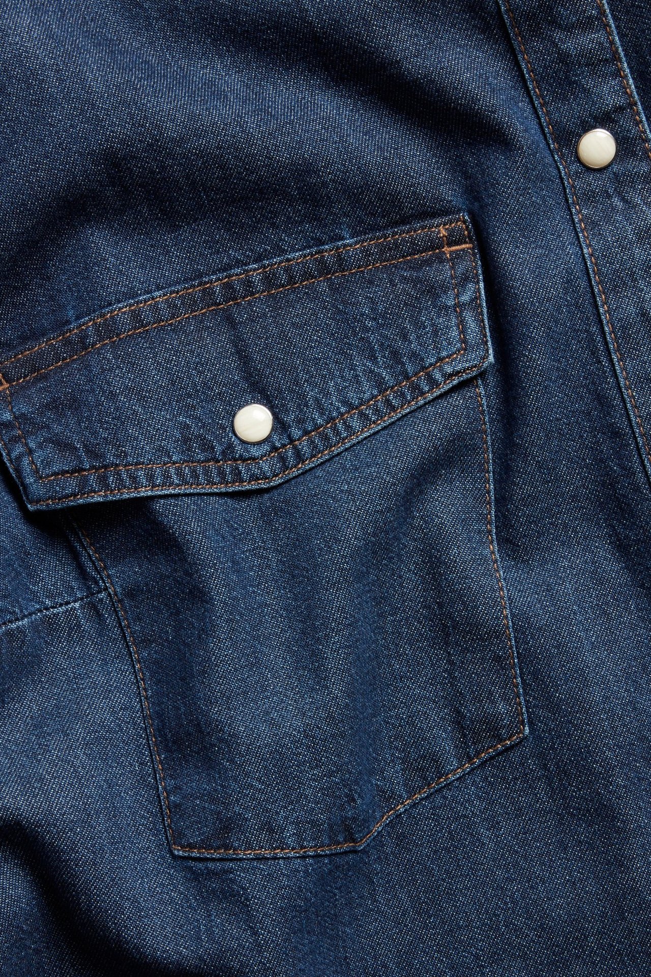 Zbliżenie na ciemnoniebieską koszulę jeansową z pomarańczowo obszytą kieszenią i białym napem.