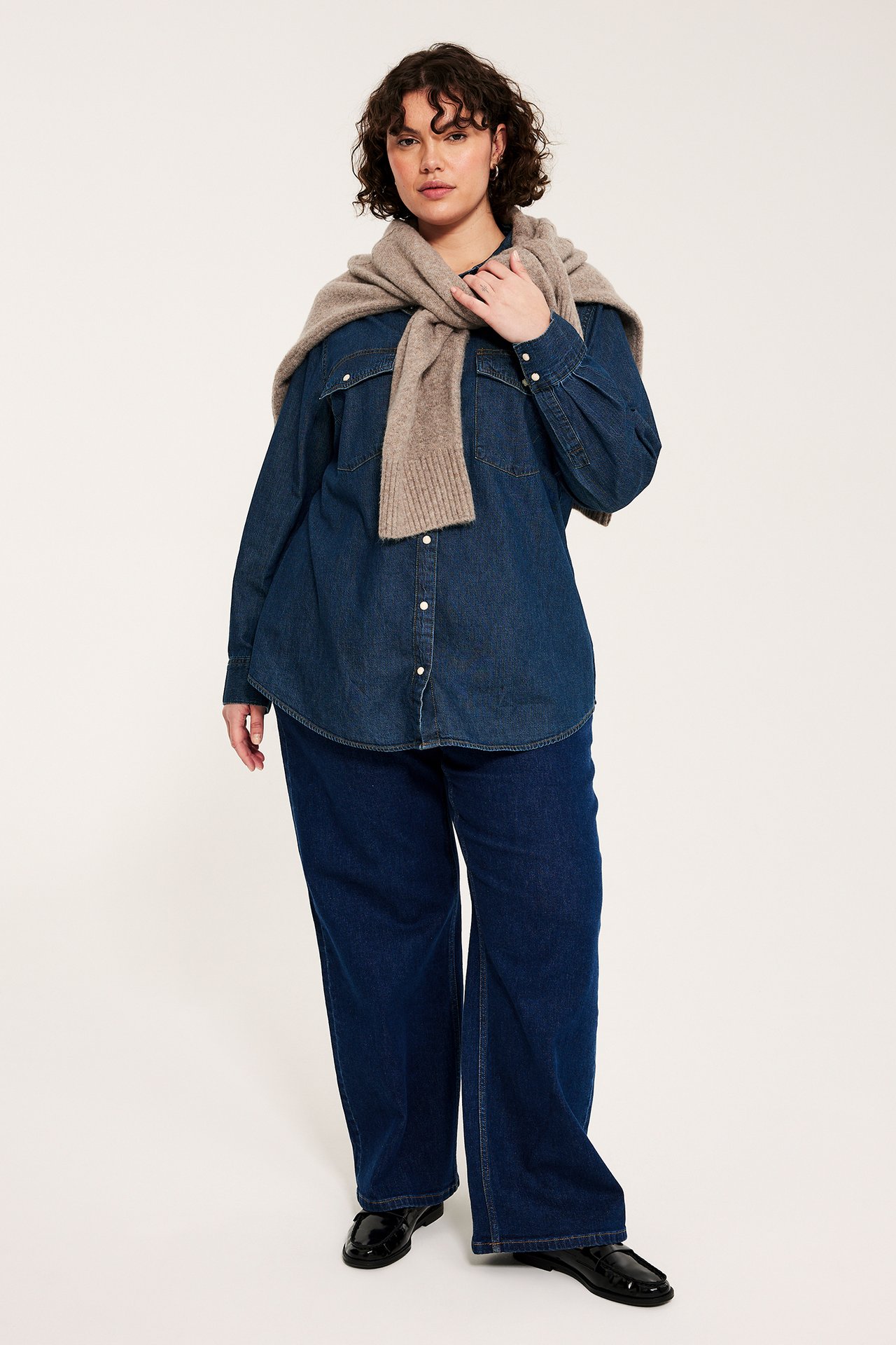 Edestä: Nainen tummansinisessä farkkupaidassa neppareilla, leveissä farkuissa, mustissa loafereissa, beige neule hartioilla.