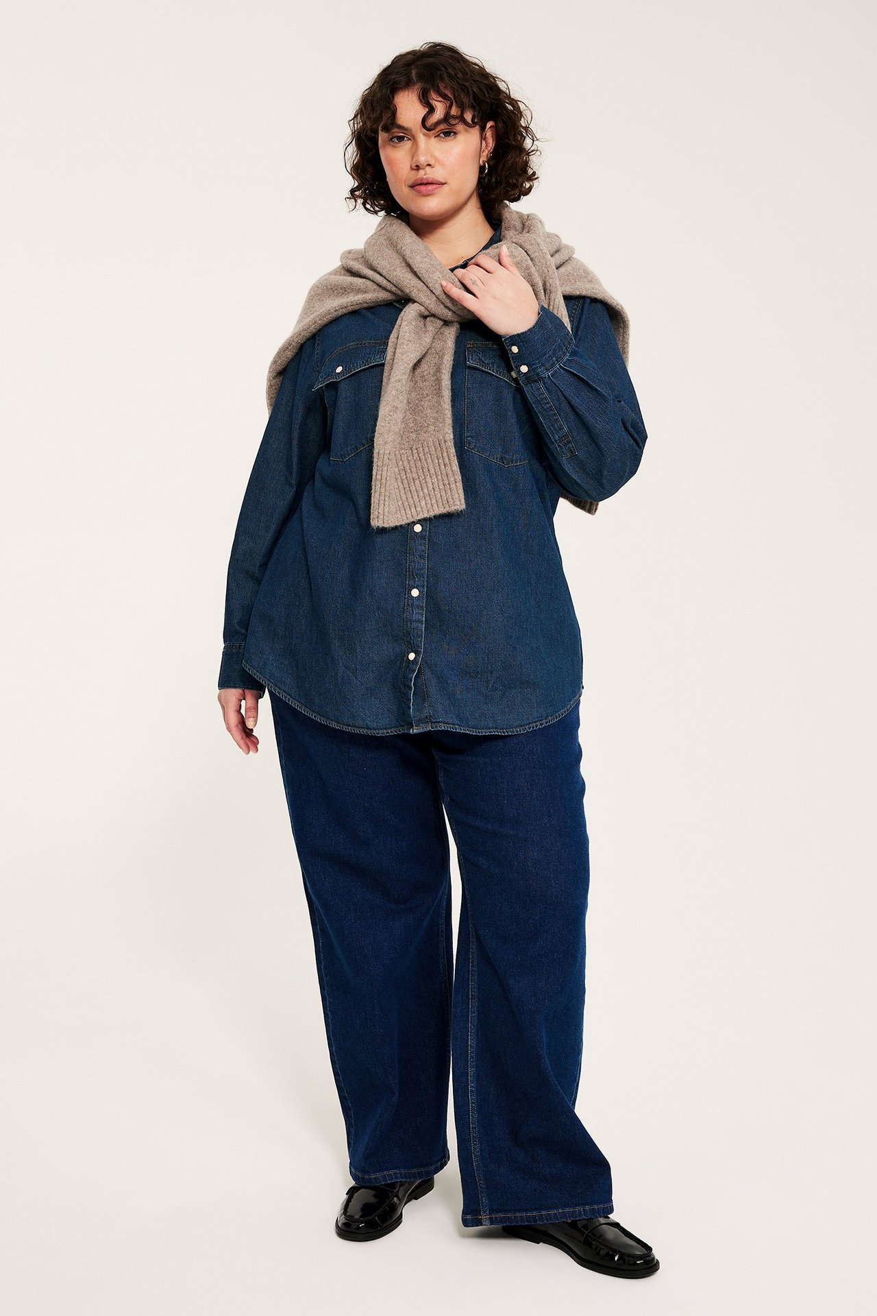 Forfra: Kvinne i mørk blå denimskjorte med trykknapper, vide jeans, svarte loafers, med en drapert beige genser.