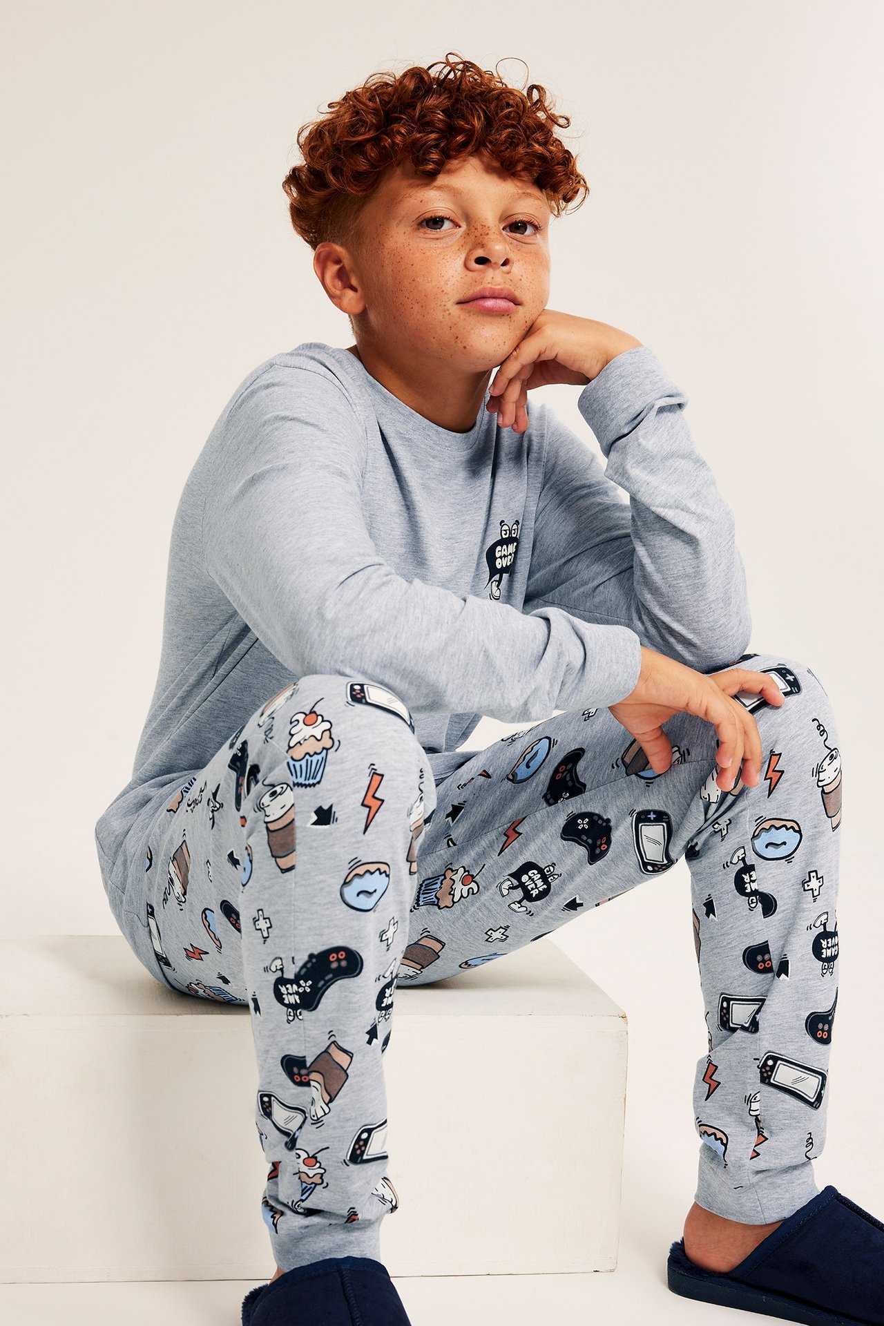Edestäpäin lapsi harmaassa pitkähihaisessa pelipainatetussa pyjamassa ja tohveleissa.