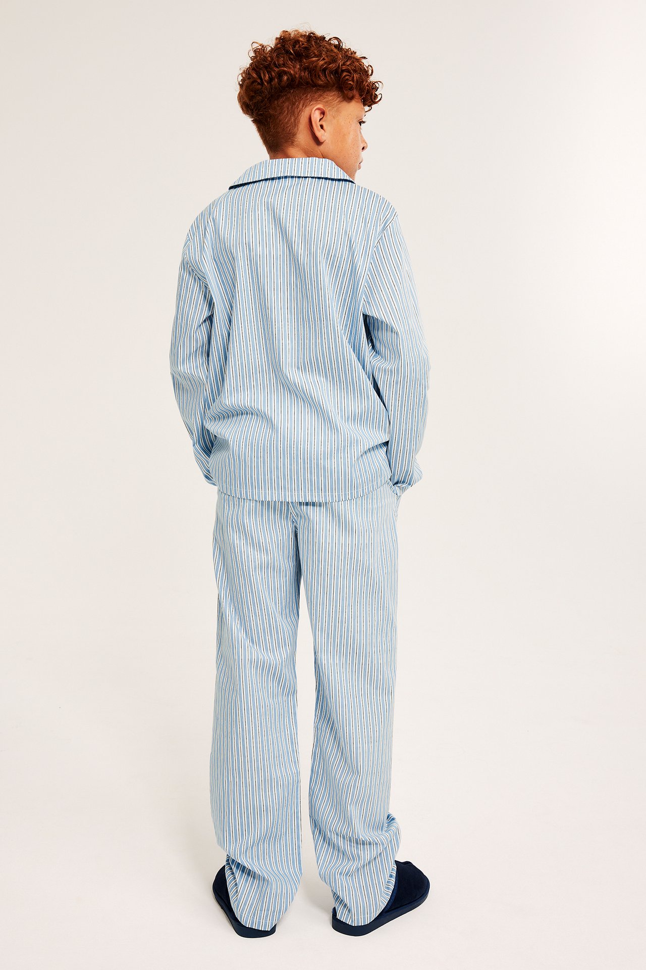 Ryggvy: barn i ljusblårandig pyjamas i poplin med krage, raka byxor och mörkblå tofflor.