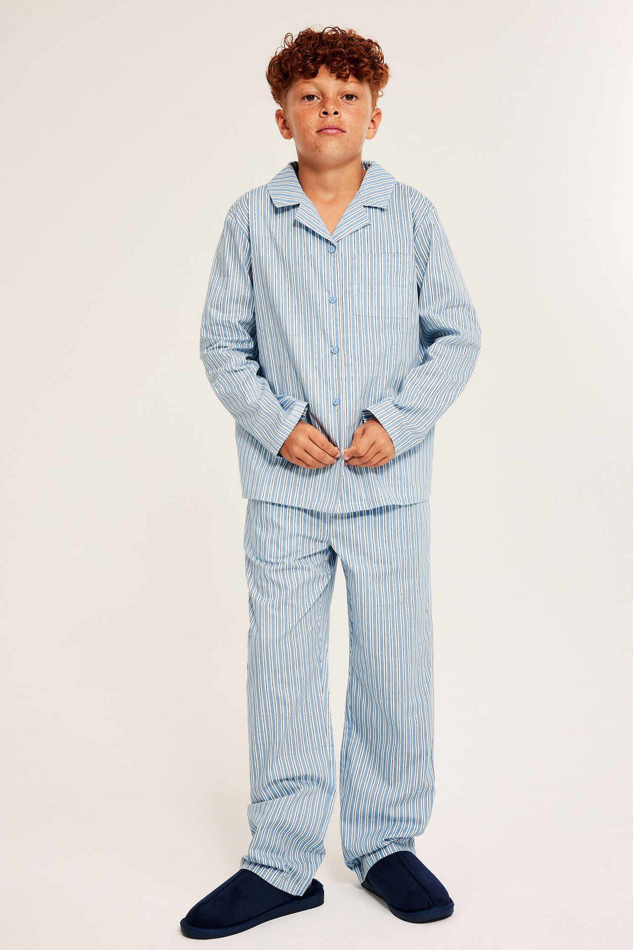 Framifrån: Kid bär en ljusblå randig pyjamas i poplin och mörkblå tofflor.