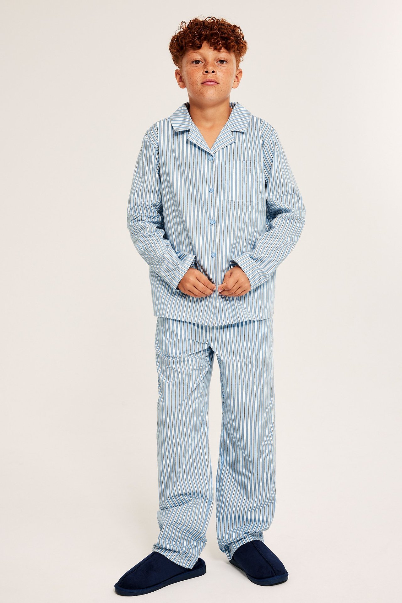 Widok z przodu: Dziecko w jasnoniebieskiej, pasiastnej piżamie z popeliny i granatowych kapciach.