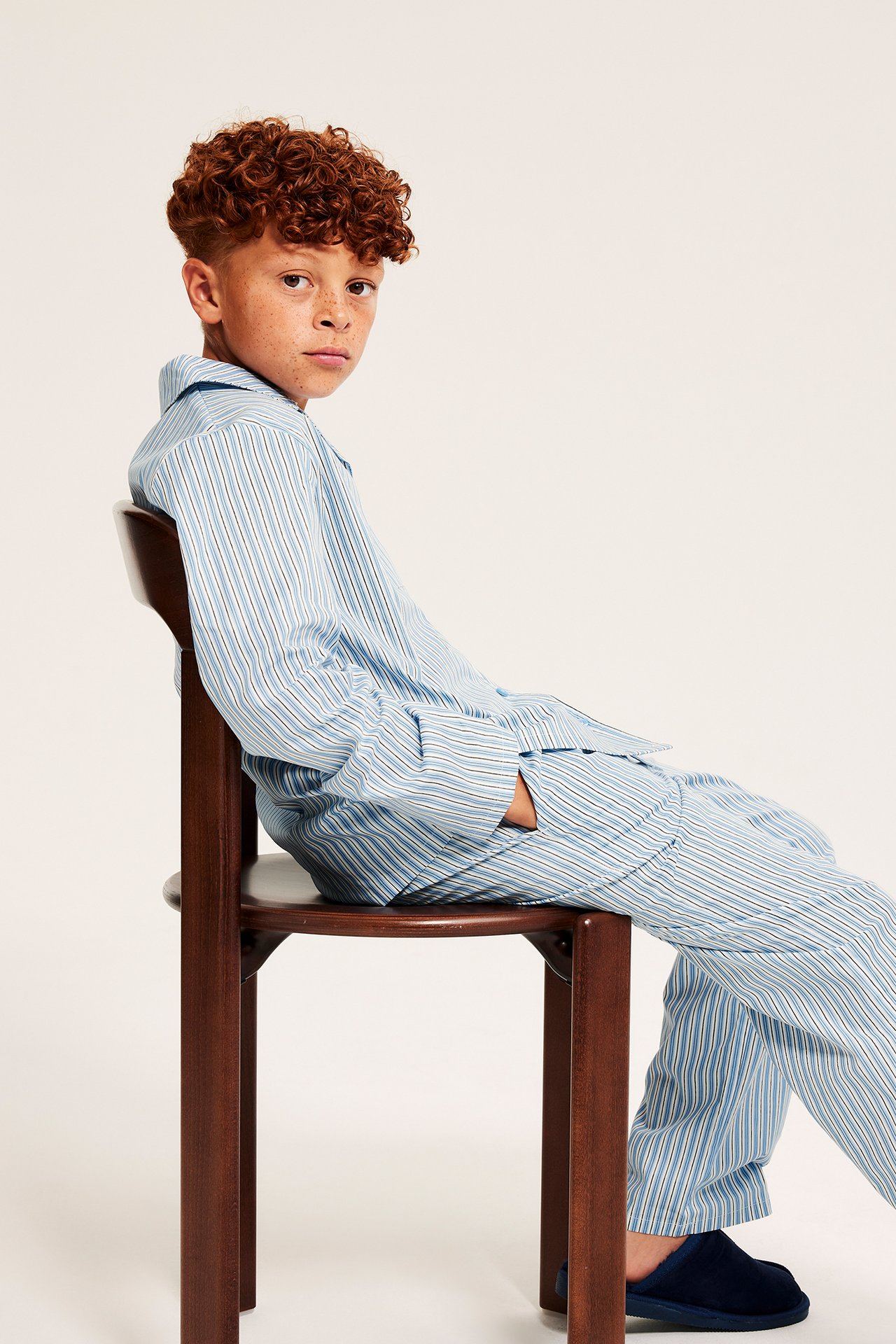 Sidovy av ett barn i ljusblå randig pyjamas i poplin med knappar, krage och mörkblå tofflor.