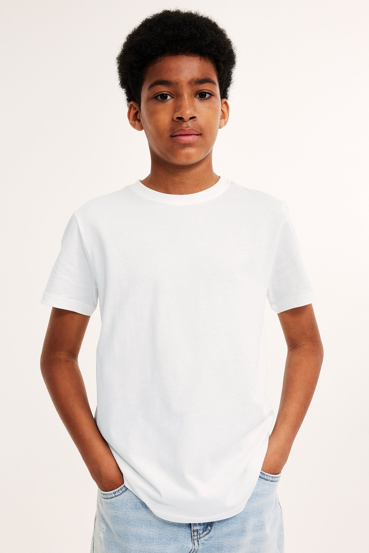 Edestä kuvattu lapsi valkoisessa puuvilla-T-paidassa ja vaaleansinisissä farkuissa, kädet taskuissa.