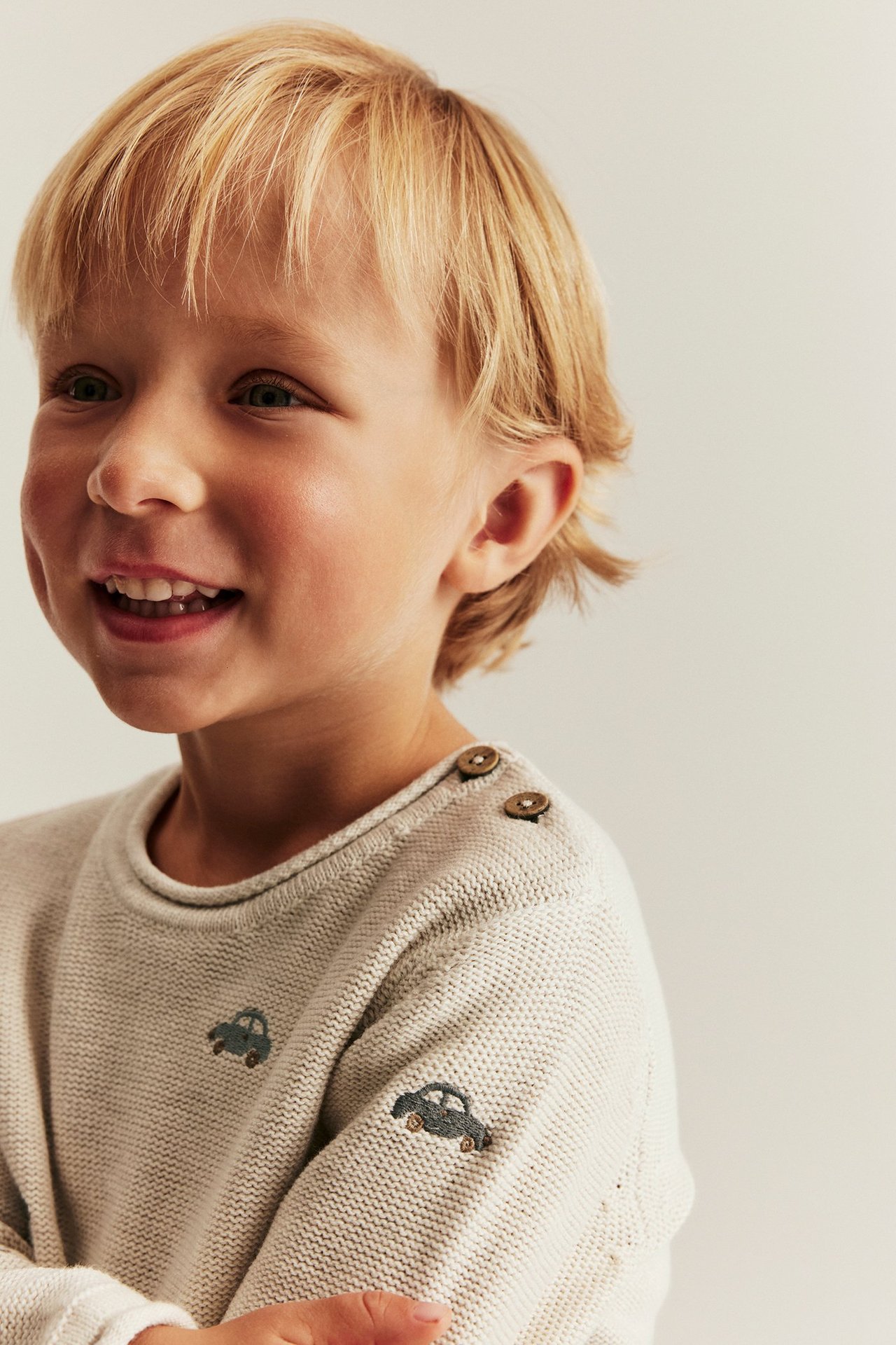 Nærbilde av et smilende barn i en beige strikket bomullsgenser med broderte biler og skulderknapper.