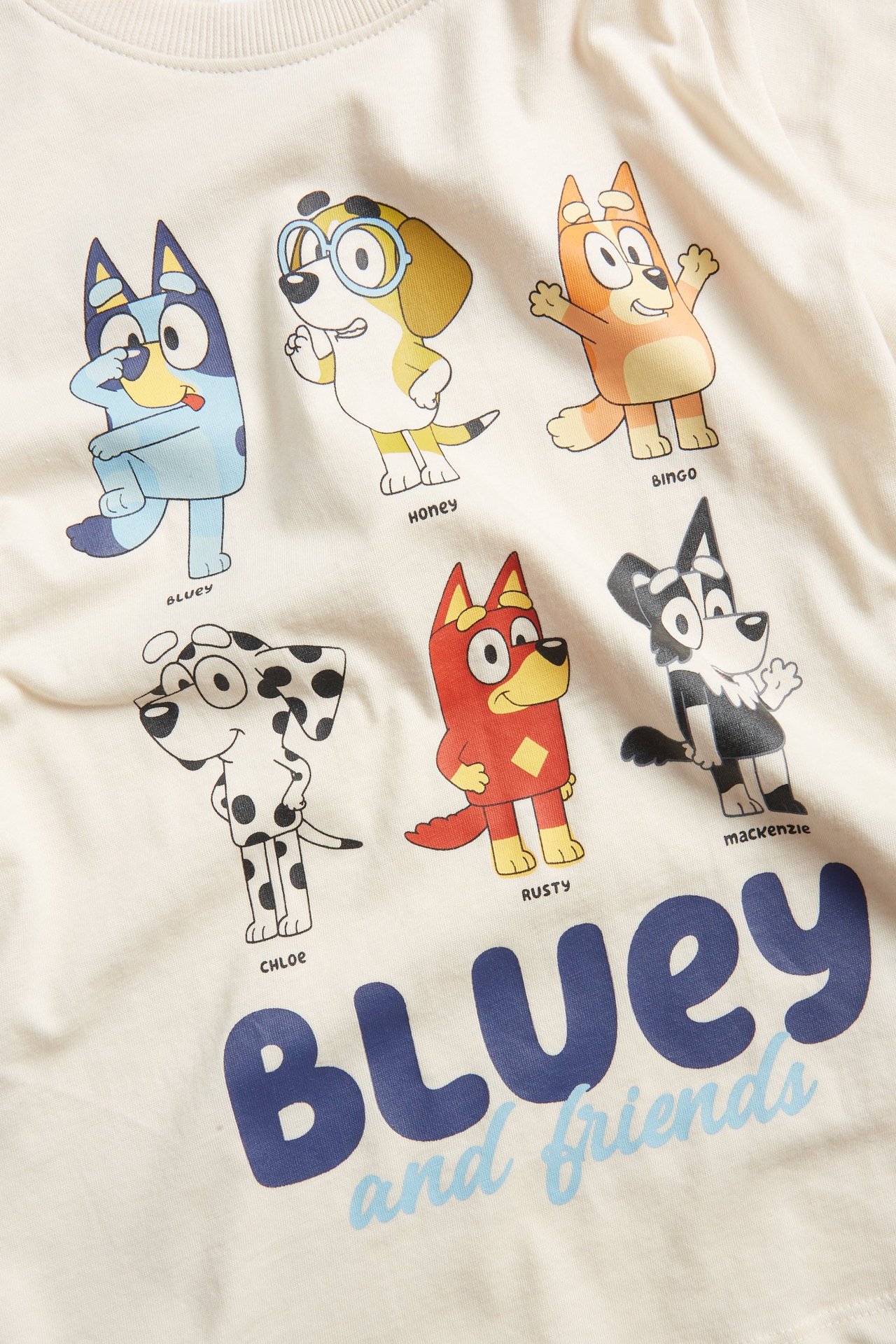 Frontvy av en ljusbeige bomulls-t-shirt för barn med stort färgglatt Bluey och vänner-tryck.