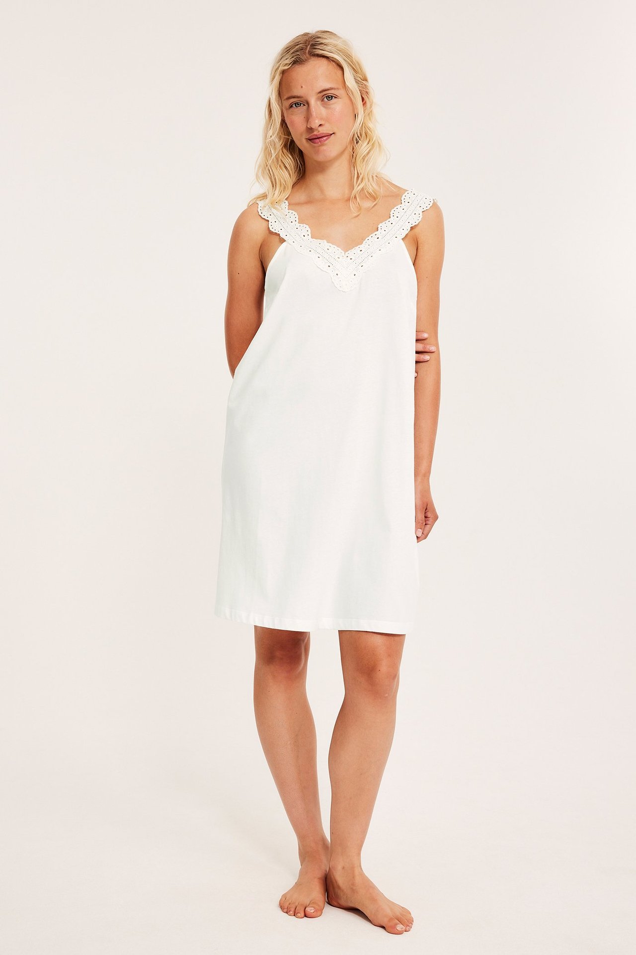 Edestä: nainen yllään luonnonvalkoinen puuvillatrikoo yömekko pitsisellä V-pääntiellä ja olkaimilla.