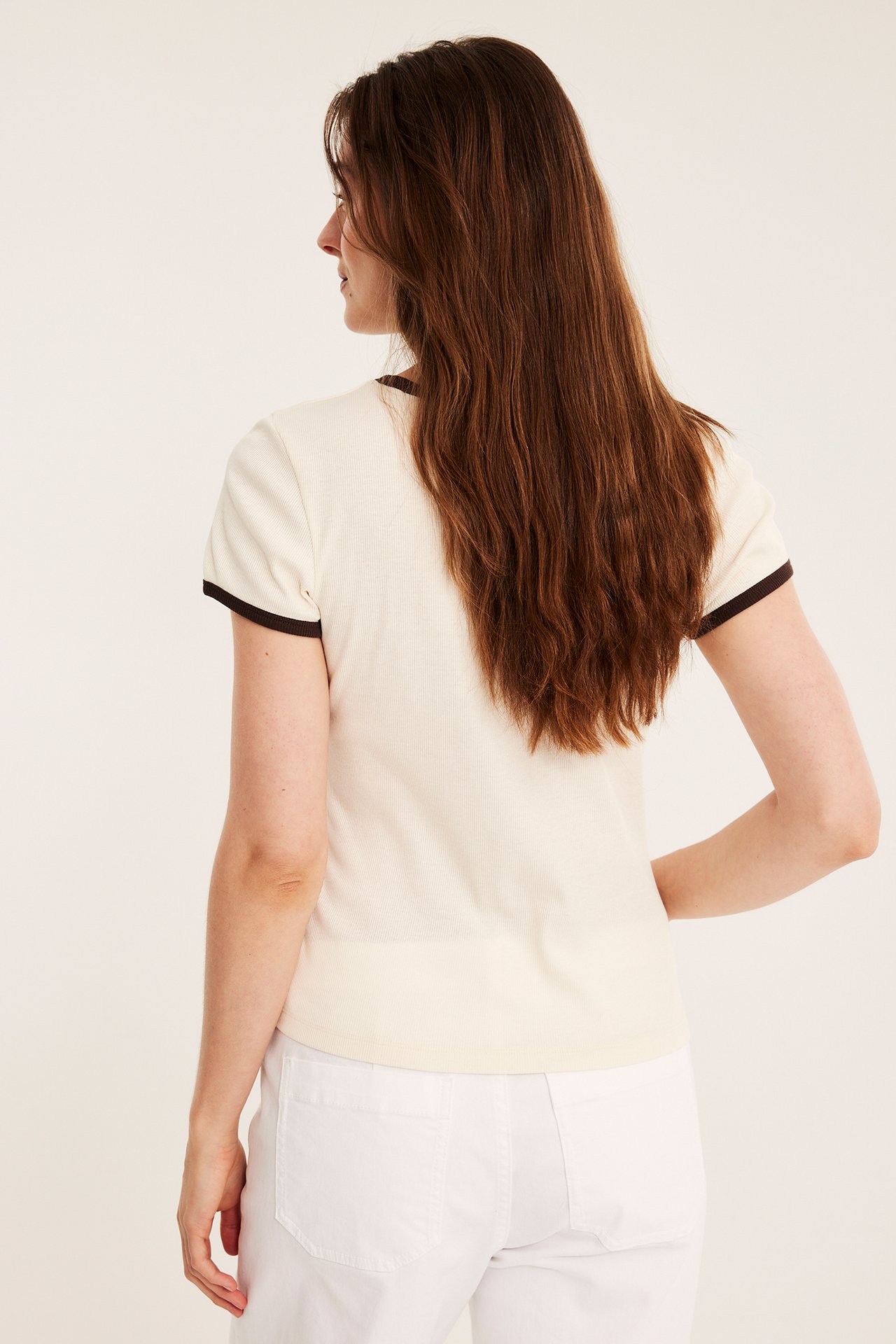 Ryggvy av kvinna i offwhite ribbad baby-t-shirt med mörka kantband och vita byxor.