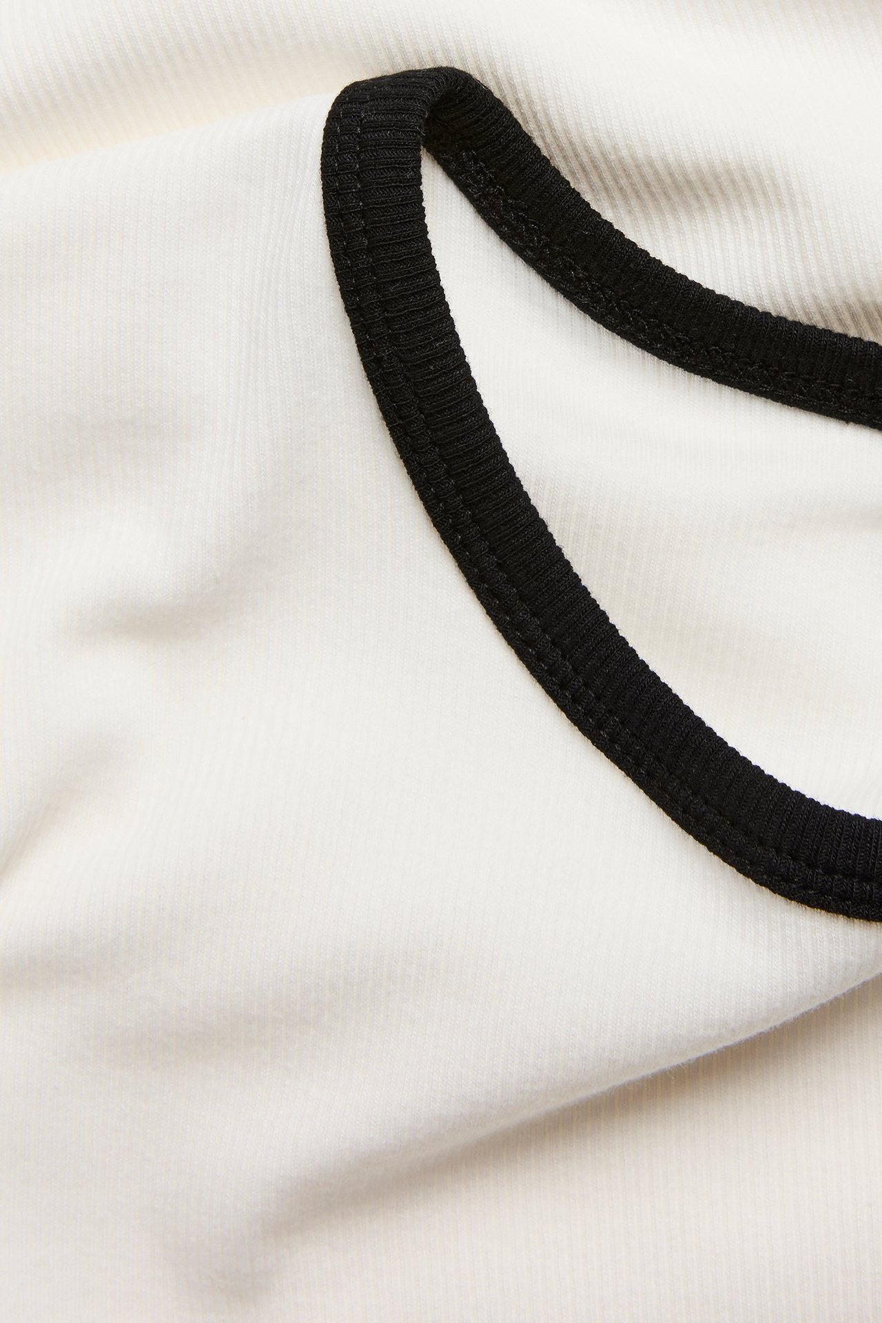 Zbliżenie na kremowy prążkowany T-shirt typu baby tee z czarnymi kontrastowymi lamówkami przy dekolcie.