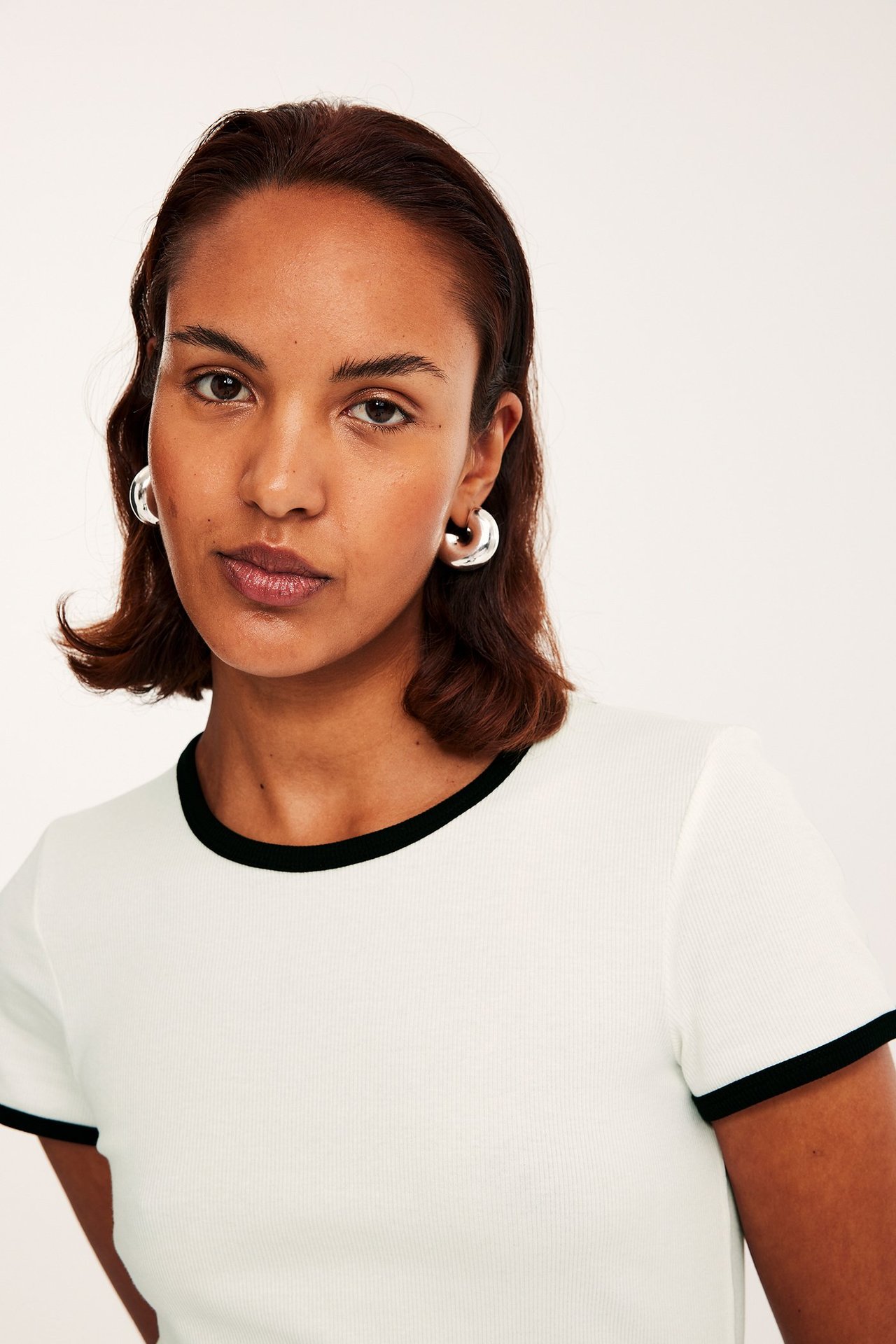 Zbliżenie kobiety w kremowym, prążkowanym topie z krótkim rękawem, czarnymi lamówkami i srebrnymi kolczykami.