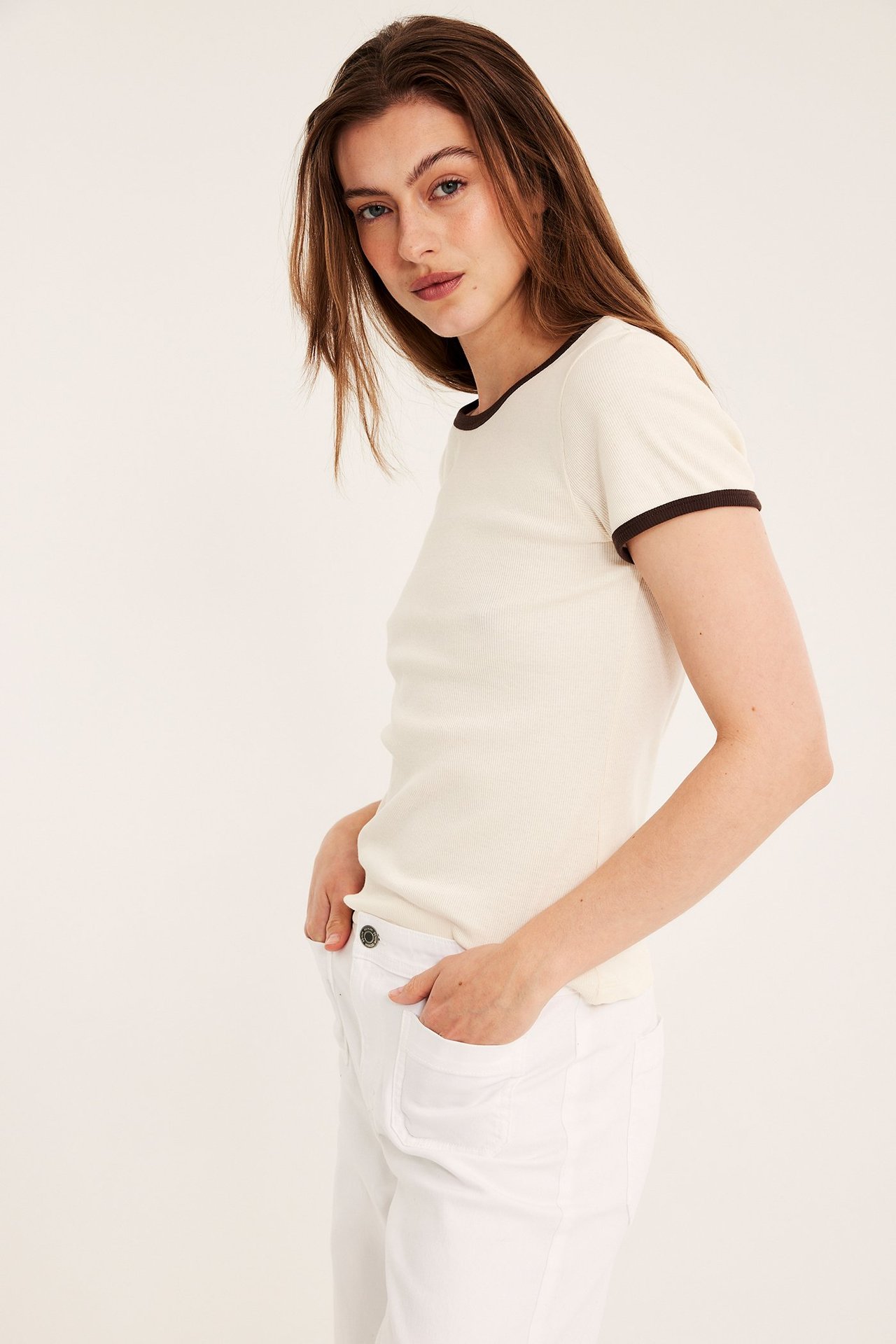 Sivukuva naisesta, jolla on luonnonvalkoinen ribbineuleinen lyhythihainen toppi tummanruskealla kanttauksella ja valkoiset housut.