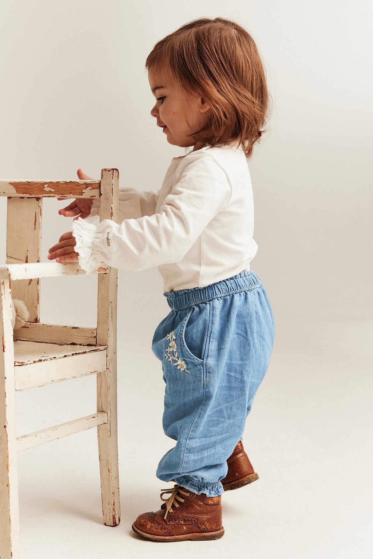 Sidevisning: baby i lyseblå broderte denimjeans, hvit topp, brune sko, holder en trestol.