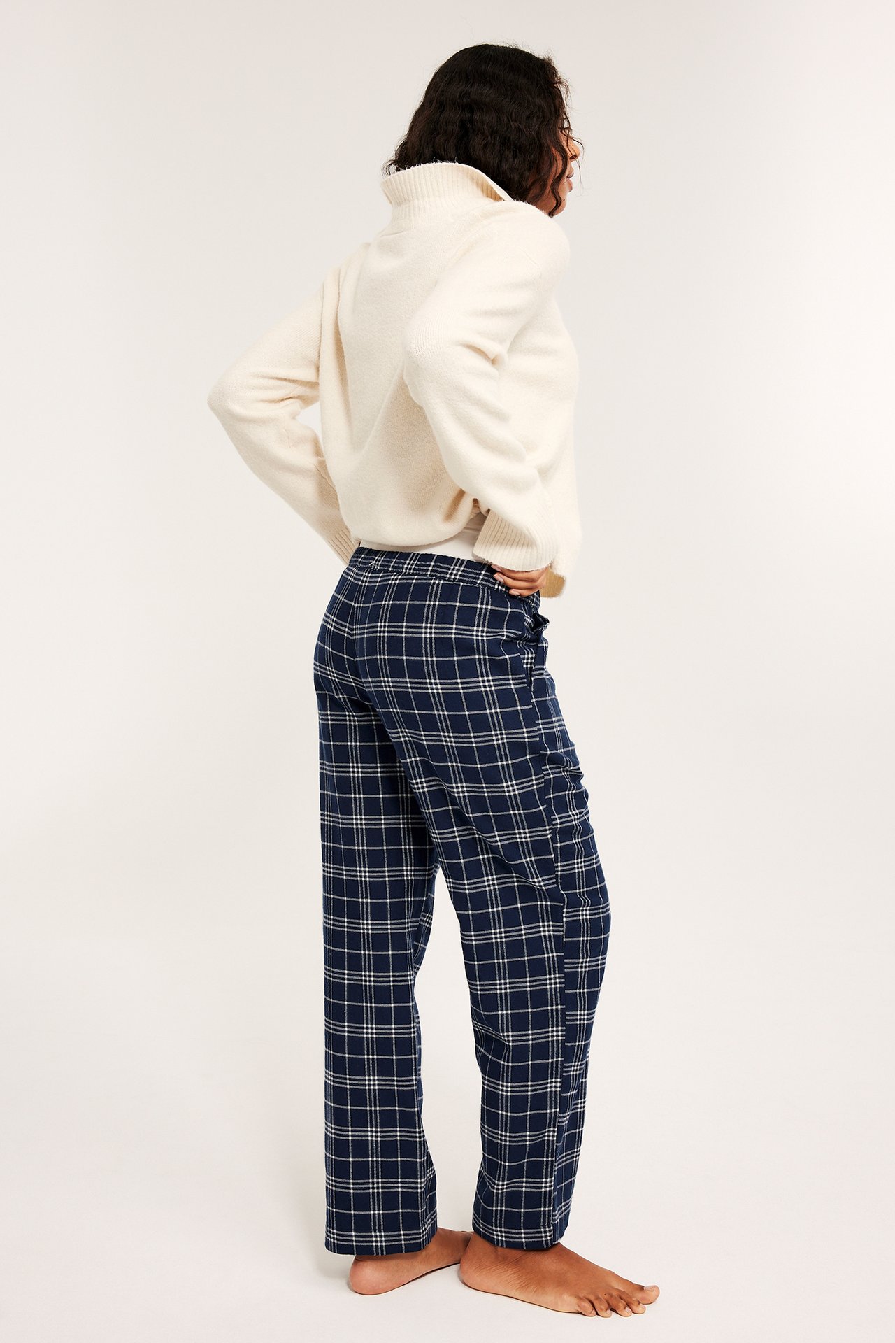 Kvinnlig person i vit stickad sweater och rutiga flanellpyjamasbyxor i mörkblått.