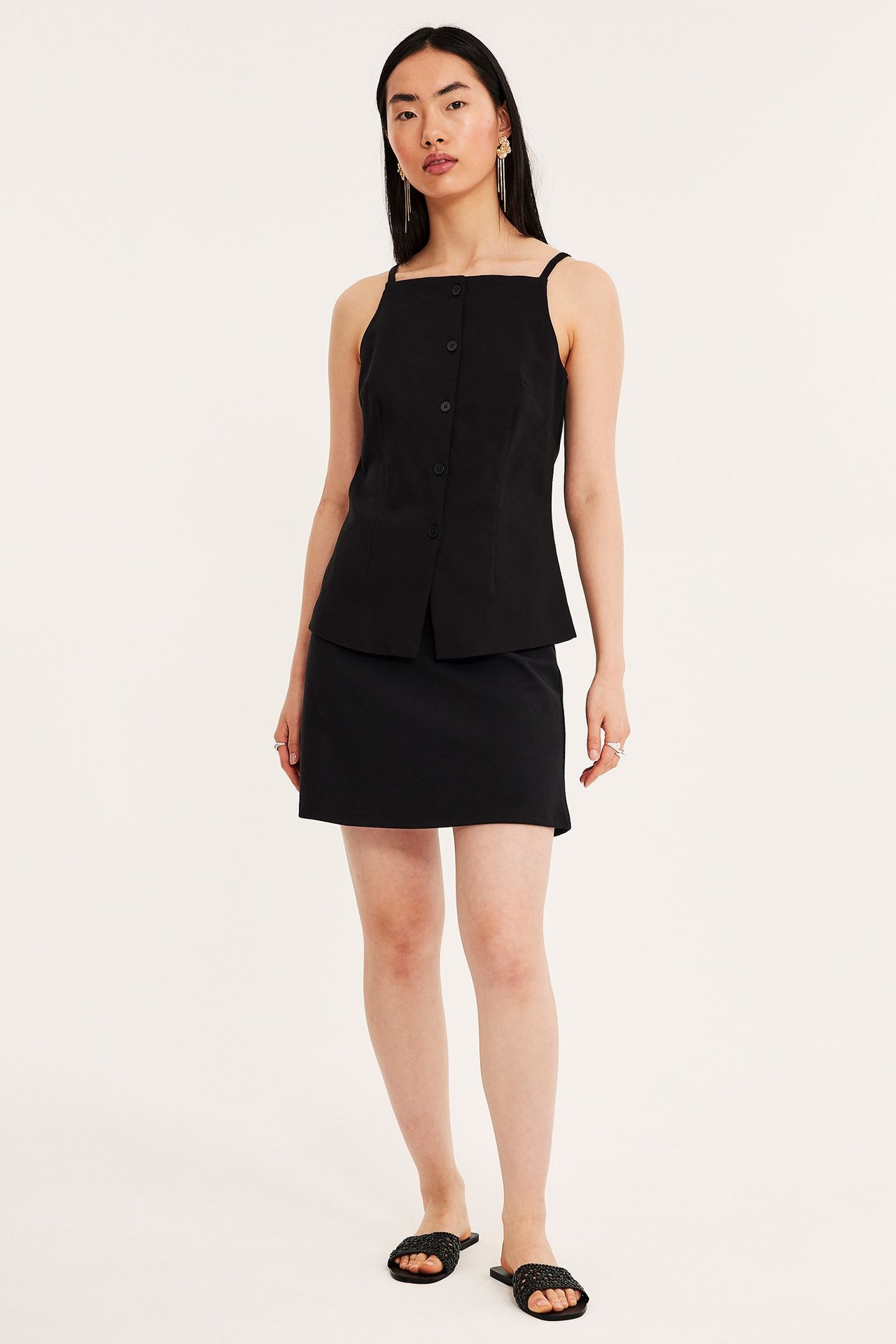Edestä kuvattu nainen mustassa A-linjaisessa minihameessa, mustassa napitetussa topissa ja mustissa sandaaleissa.
