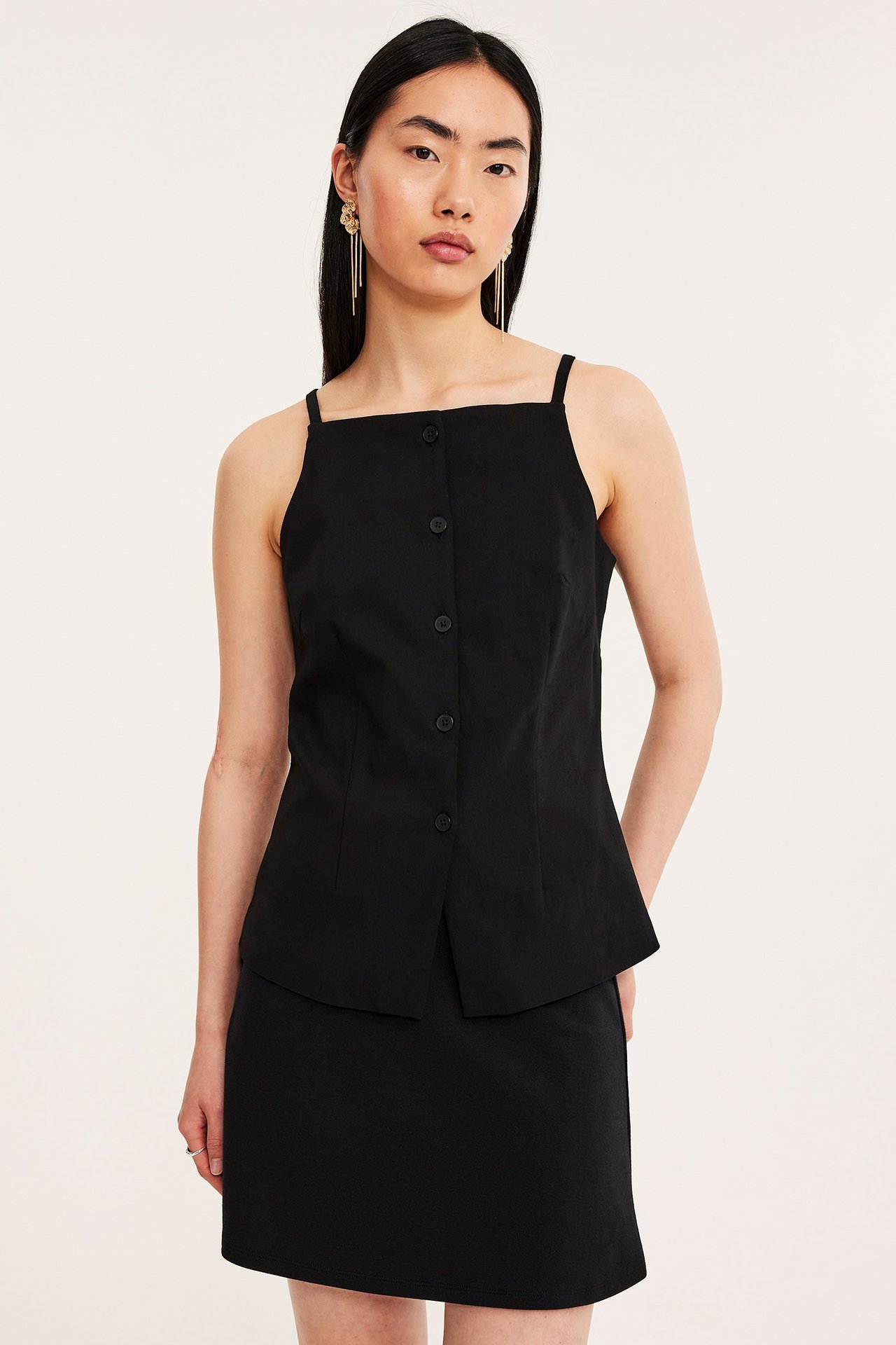 Widok z przodu: kobieta w czarnej spódnicy mini, czarnym topie na guziki i długich złotych kolczykach.