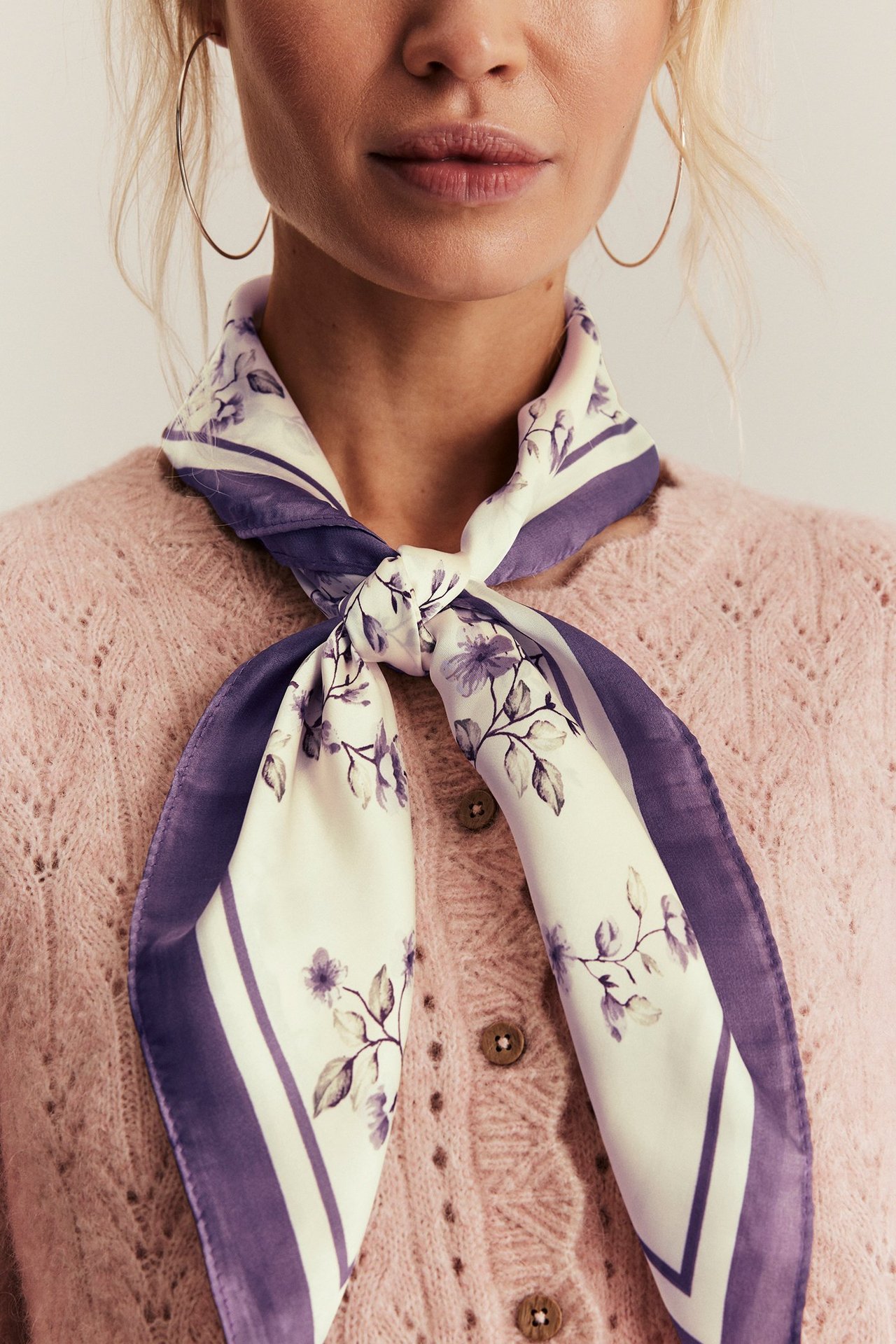 Närbild: Kvinna bär ljuslila blommig polyestersjal knuten, guldhoopörhängen och en rosa kofta.