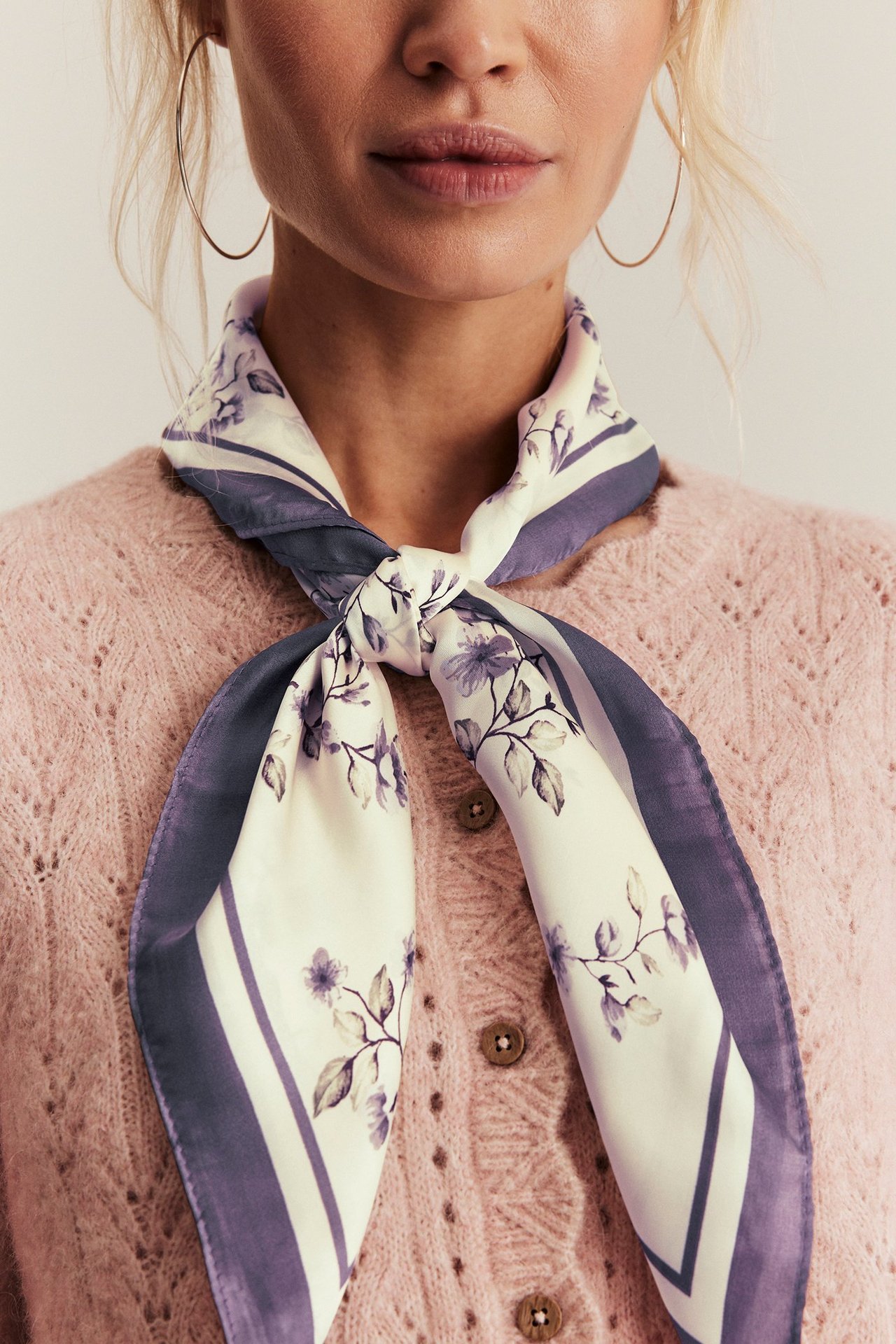 Forfra: kvinne i lys lilla blomstret polyesterskjerf knyttet over rosa strikket cardigan, med gylne ringøredobber.